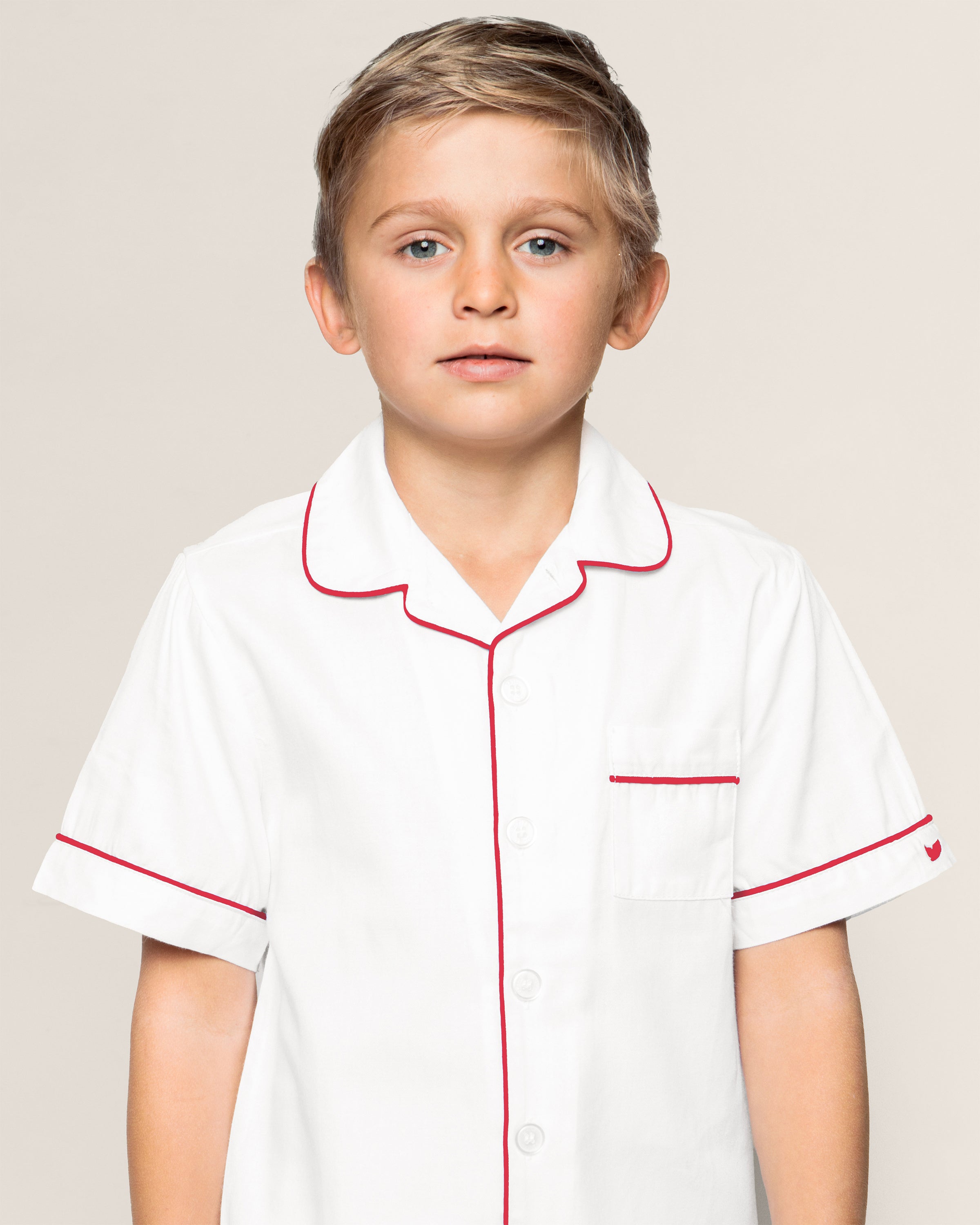 A young boy models Petite Plumes Kids White Twill Short Set with Red Piping. The yarn-dyed cotton set features a button-down shirt with red piping and a pocket on the left side. His short, light-colored hair complements his direct gaze against the plain backdrop.