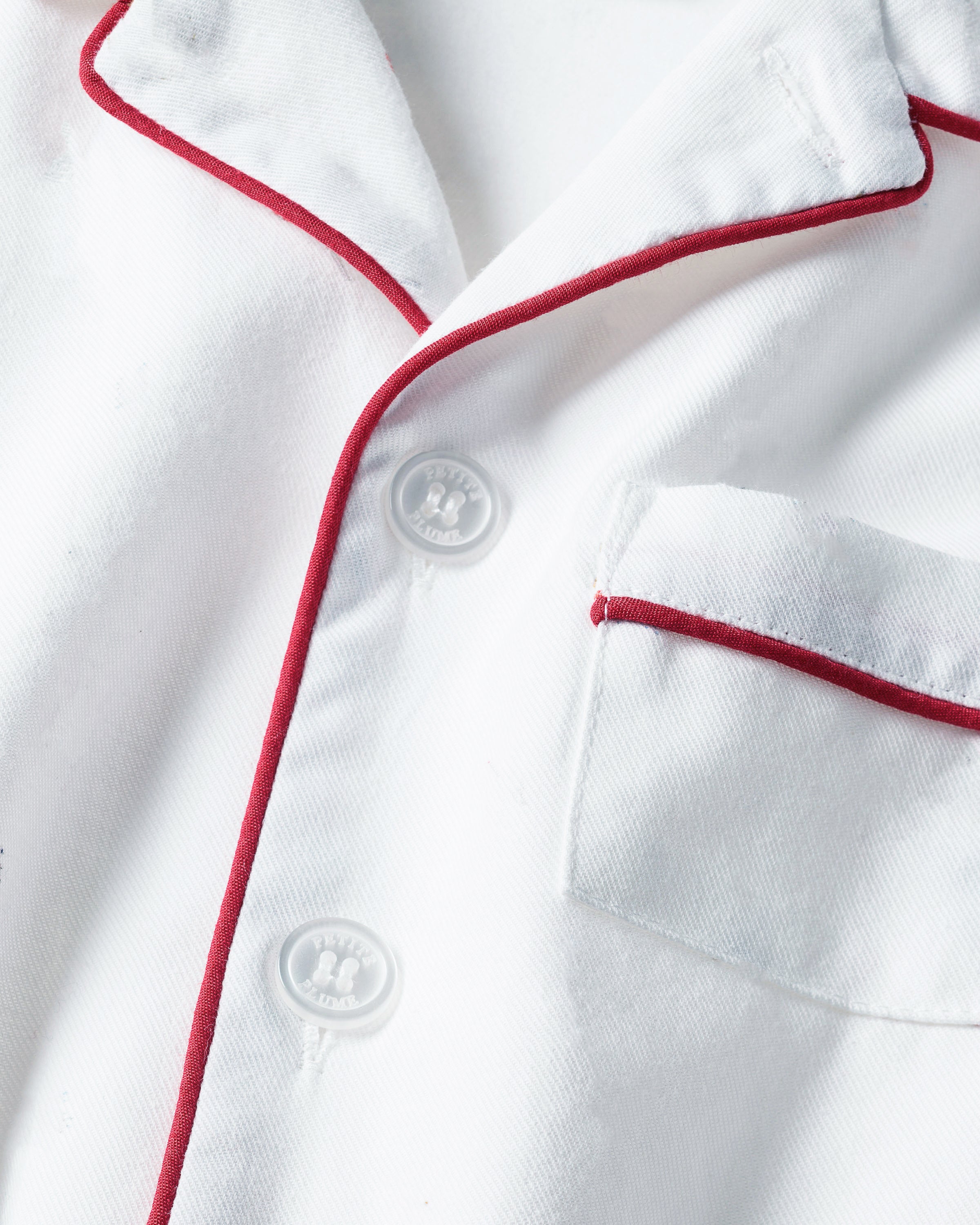 A close-up of Petite Plumes Kids White Twill Short Set showcases a white shirt with red piping on the collar and left breast pocket. Made from yarn-dyed cotton, it features round white buttons and soft, smooth fabric.