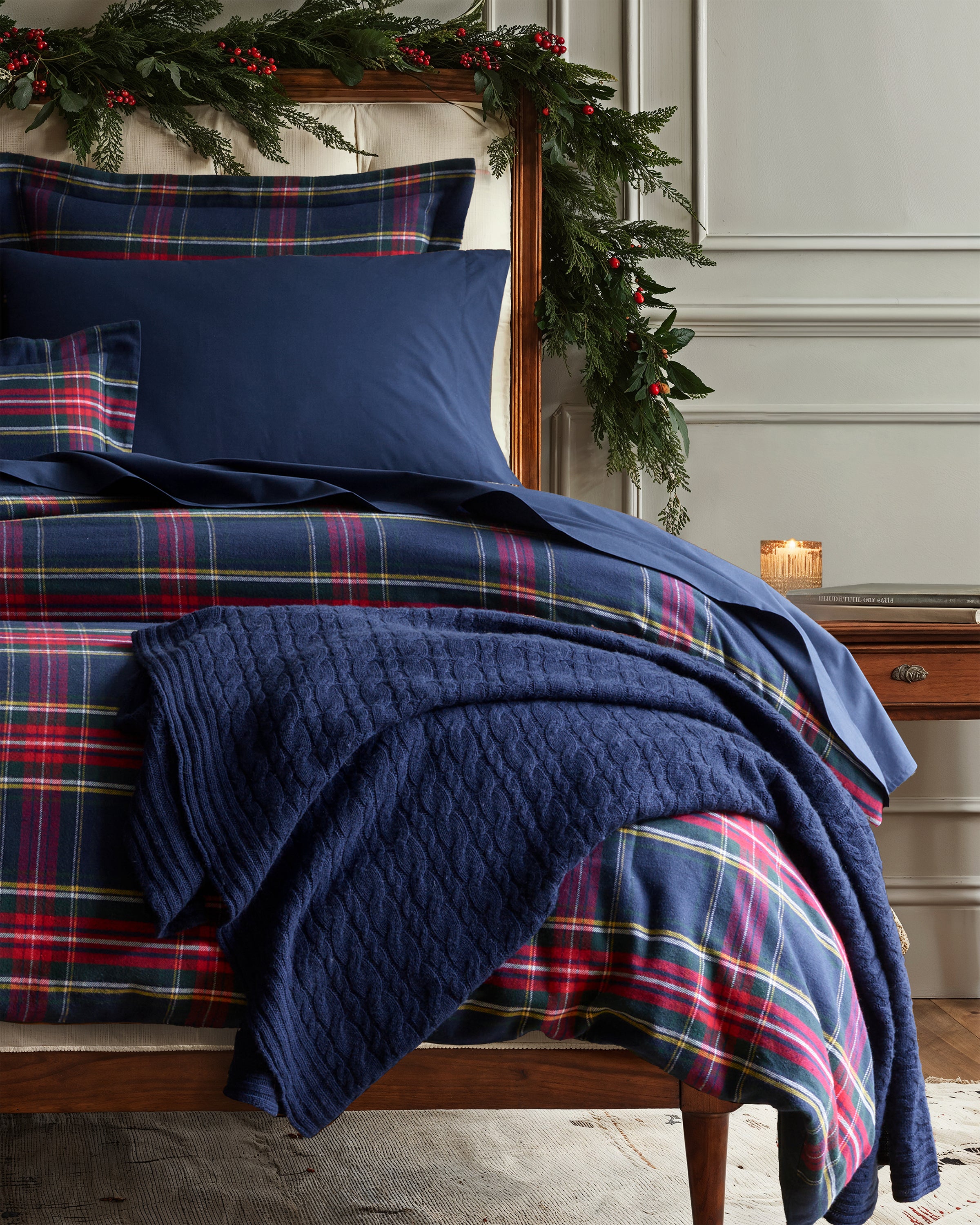 A cozy bedroom features a bed with blue and red plaid bedding, a Cashmere Cable Knit Throw in Navy by Petite Plume draped over the corner. Evergreen garlands with red berries adorn the headboard. Nearby, a lit candle on a wooden table enhances this charming giftable decor idea.