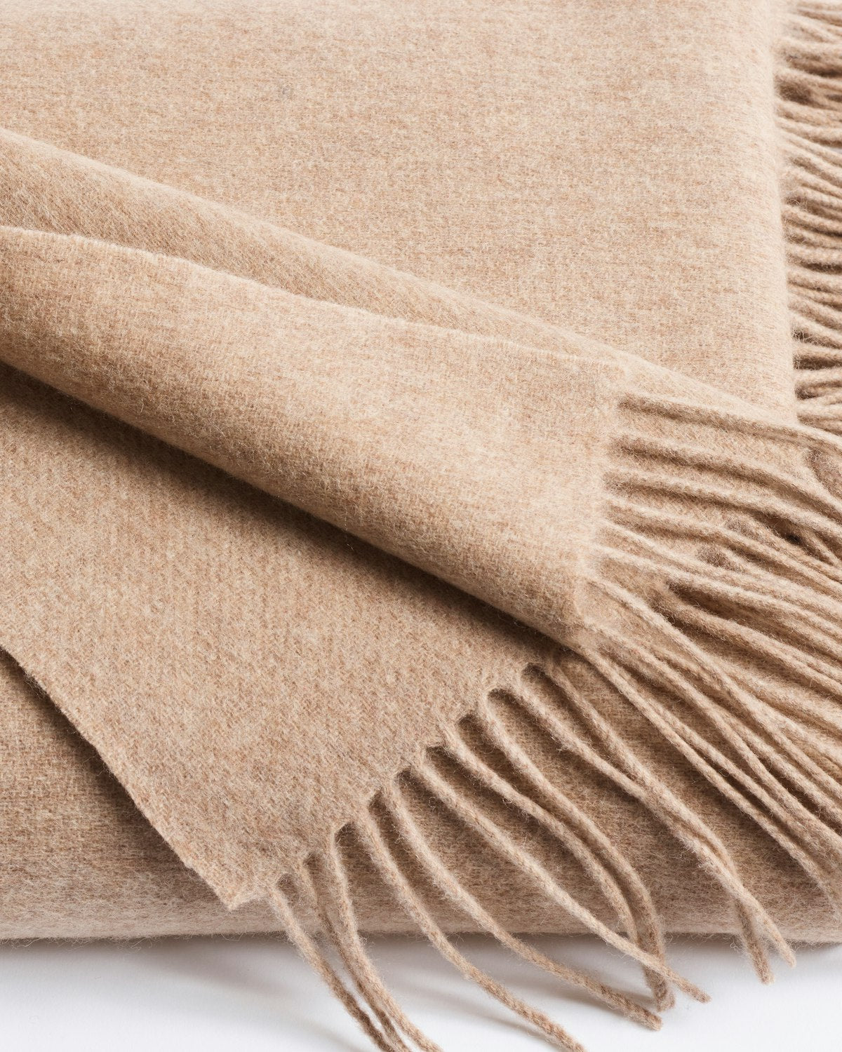 The close-up image captures a neatly folded Petite Plume Cashmere Fringe Throw in Beige. Its soft texture, fringed edges, and warm appearance make it an inviting piece of home decor.