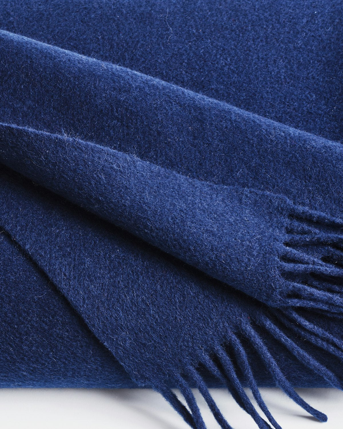 The folded Cashmere Fringe Throw in Navy by Petite Plume is a dark blue cashmere-wool blanket with fringed edges. Its soft and plush texture makes it perfect for home decor or gifting.