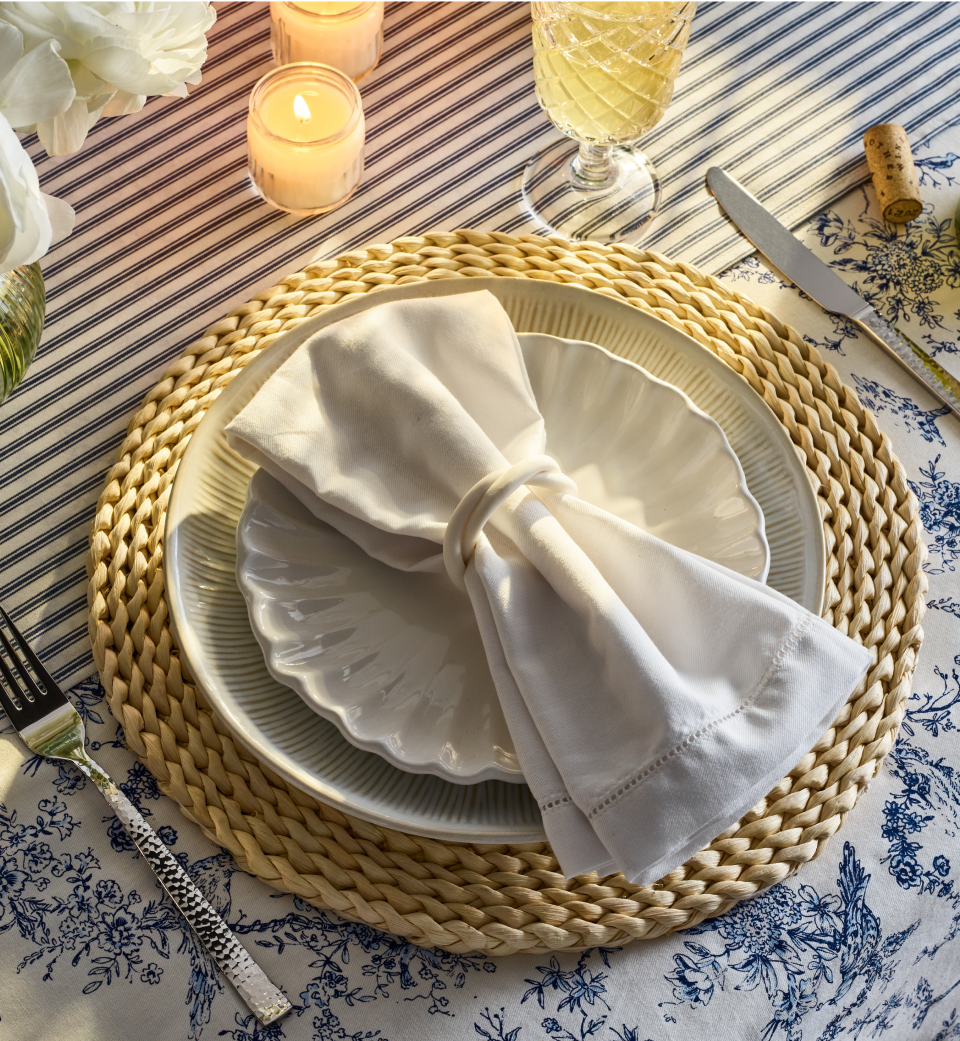 A table setting with a woven placemat, two stacked white plates, a white napkin in a ring, a fork, knife, glass of beverage, candles, and floral-patterned tablecloth in blue and white.