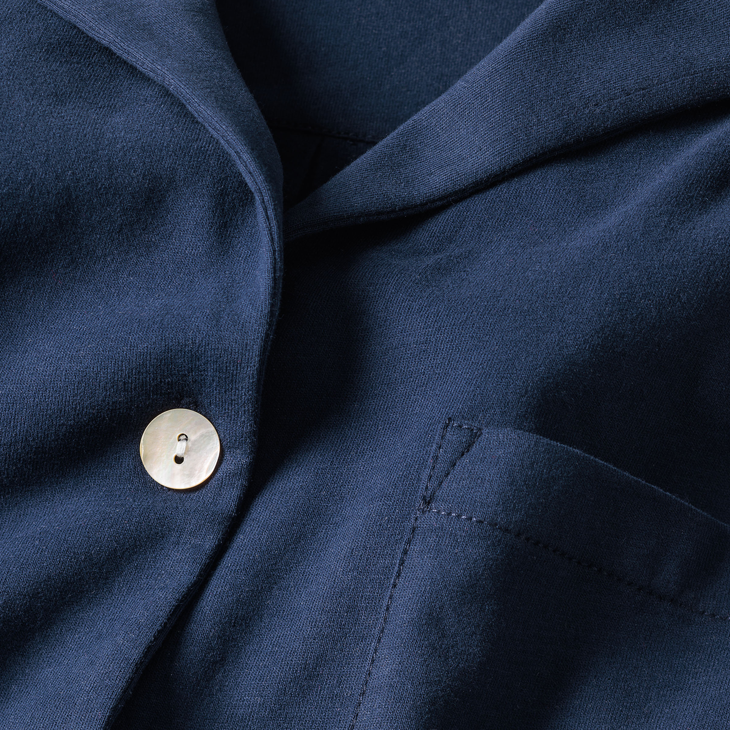 Close-up of a navy blue fabric shirt with a white button and a front pocket, showing the collar and part of the chest area. The fabric appears smooth and neatly stitched.