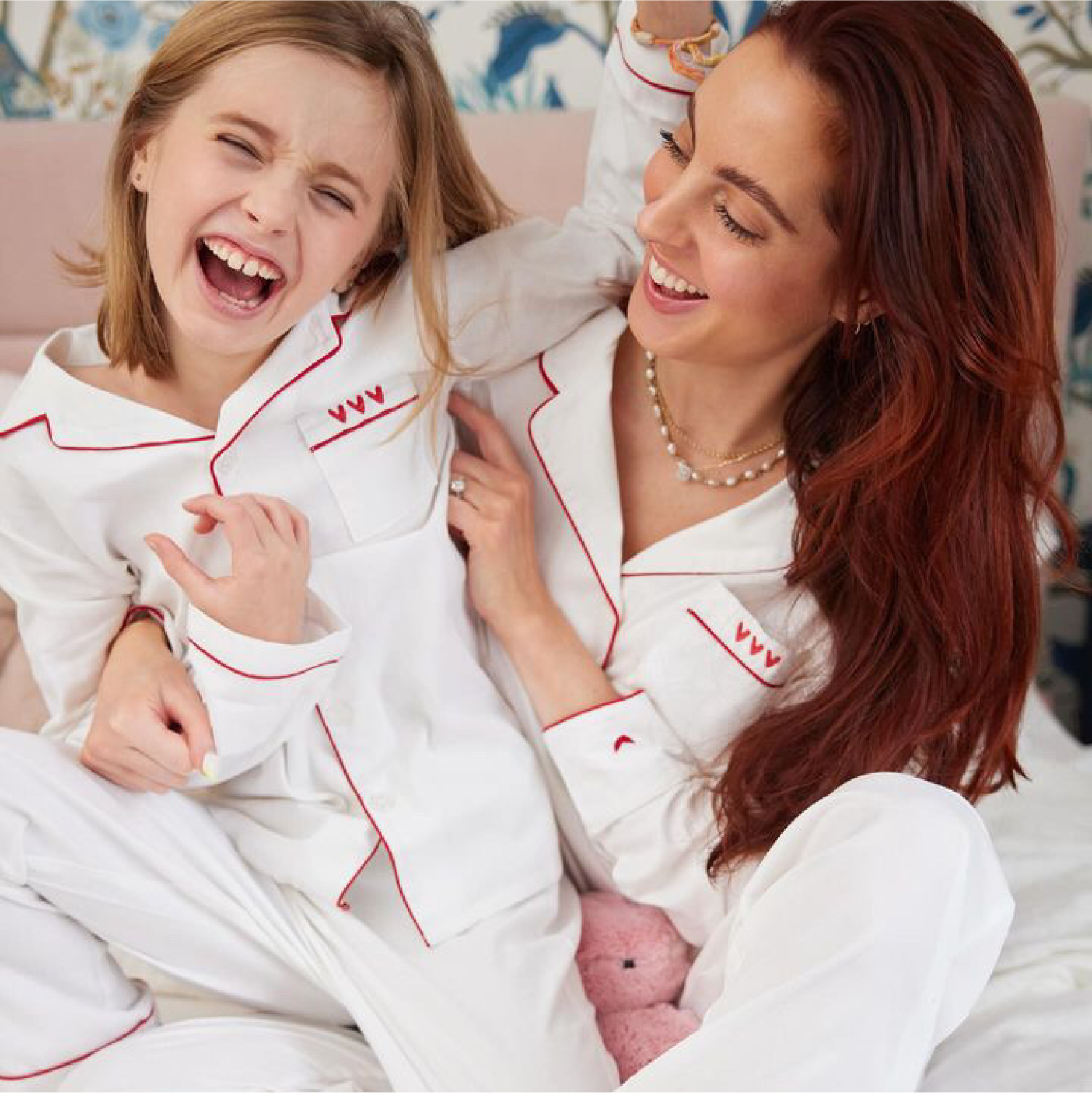 A smiling woman and a laughing young girl sit on a bed wearing matching white pajamas with red trim, enjoying a playful moment together. The background features floral wallpaper.