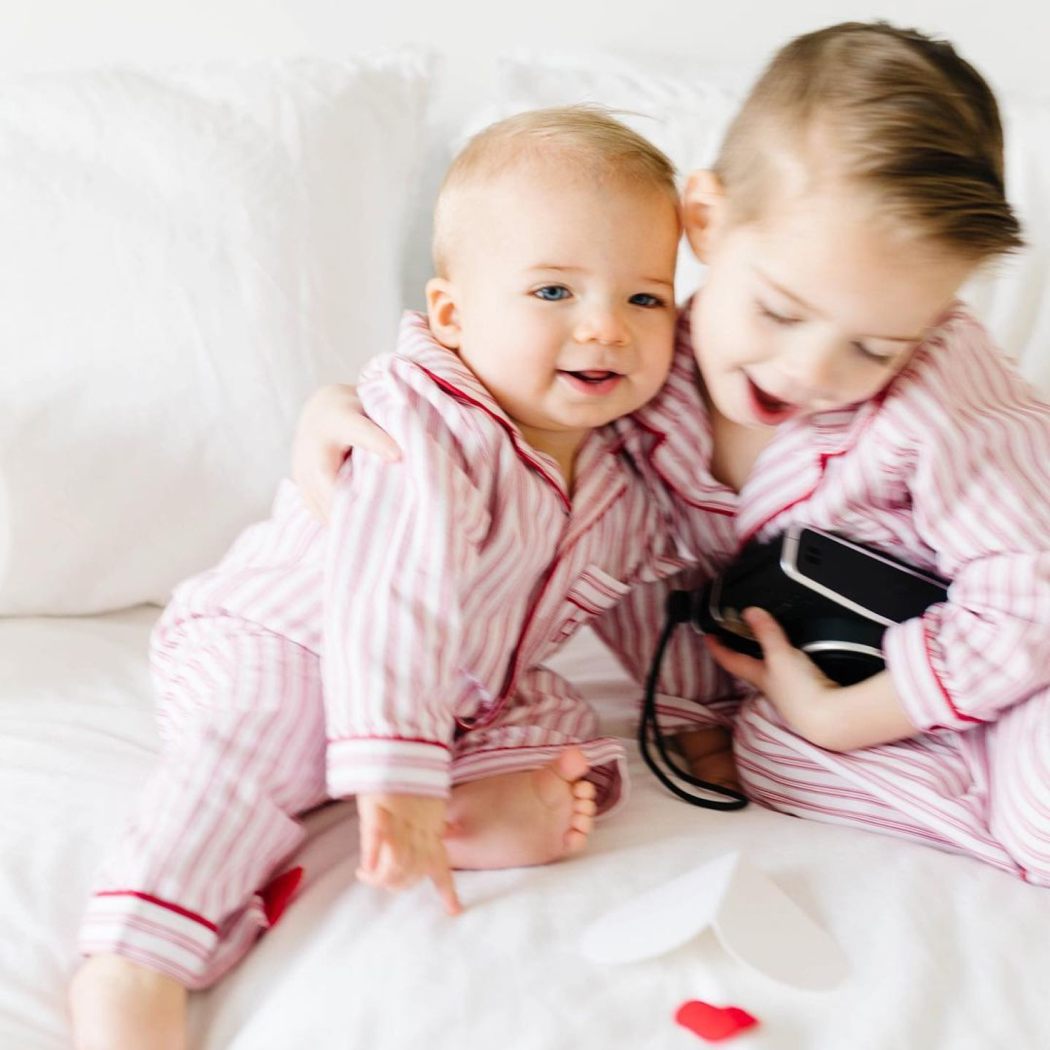 Two young children in matching pink-and-white striped pajamas sit on a bed. One child hugs the other while holding a camera. Paper hearts are scattered on the white bedding, creating a playful and loving atmosphere.