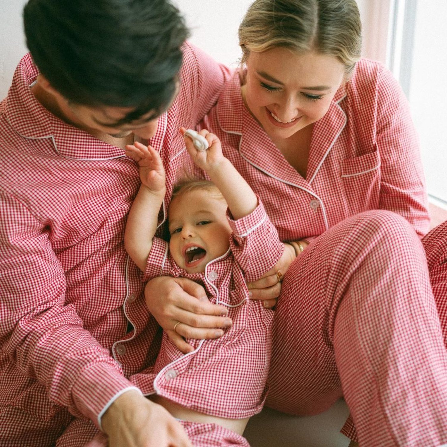 A family of three, two adults and a child, sit close together wearing matching red and white checkered pajamas. The adults smile, while the child laughs joyfully with arms raised.