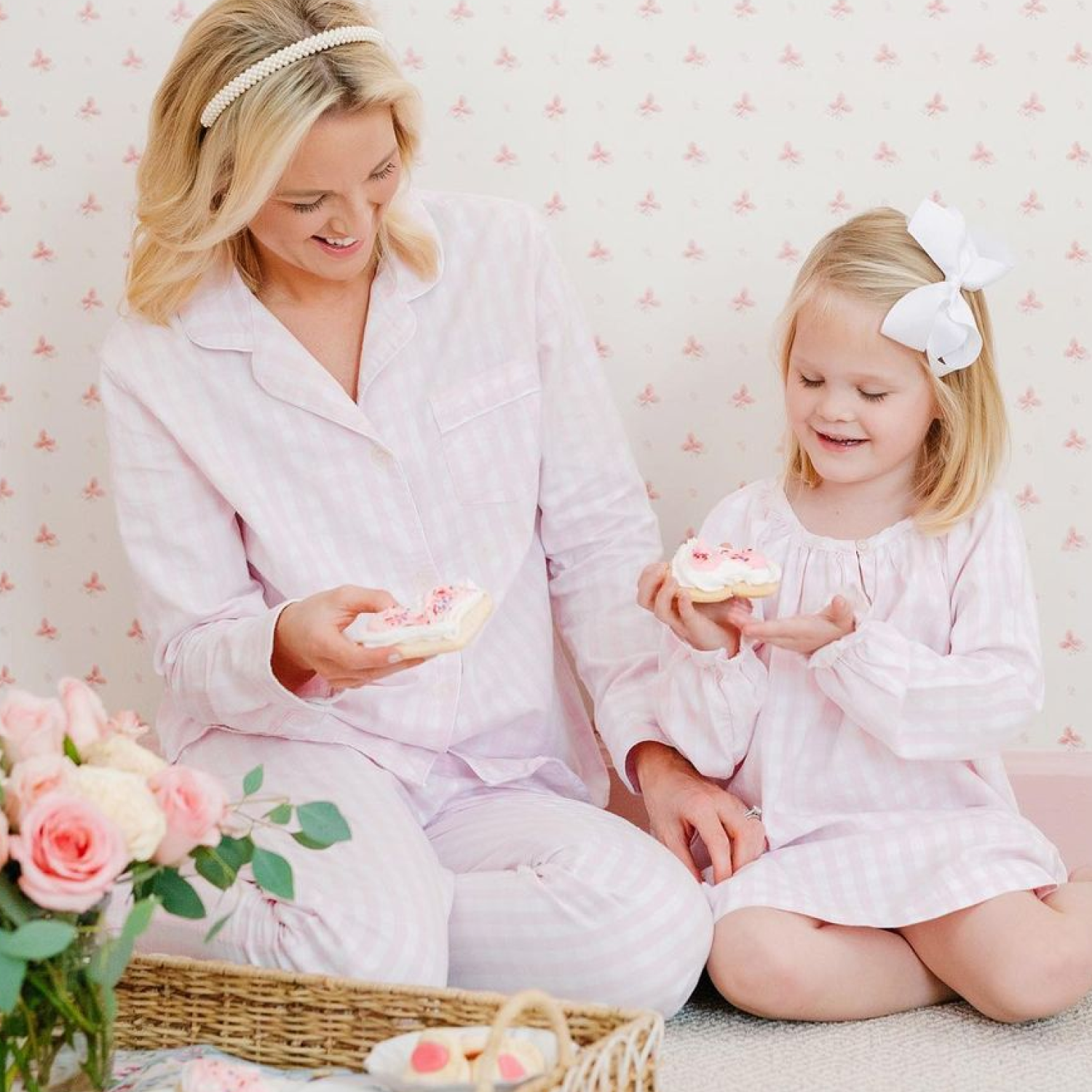 A woman and a young girl, both in matching pastel pink striped pajamas, sit on the floor smiling and holding frosted cookies. A basket with flowers and eggs is in front of them. The background is light with a faint floral pattern.