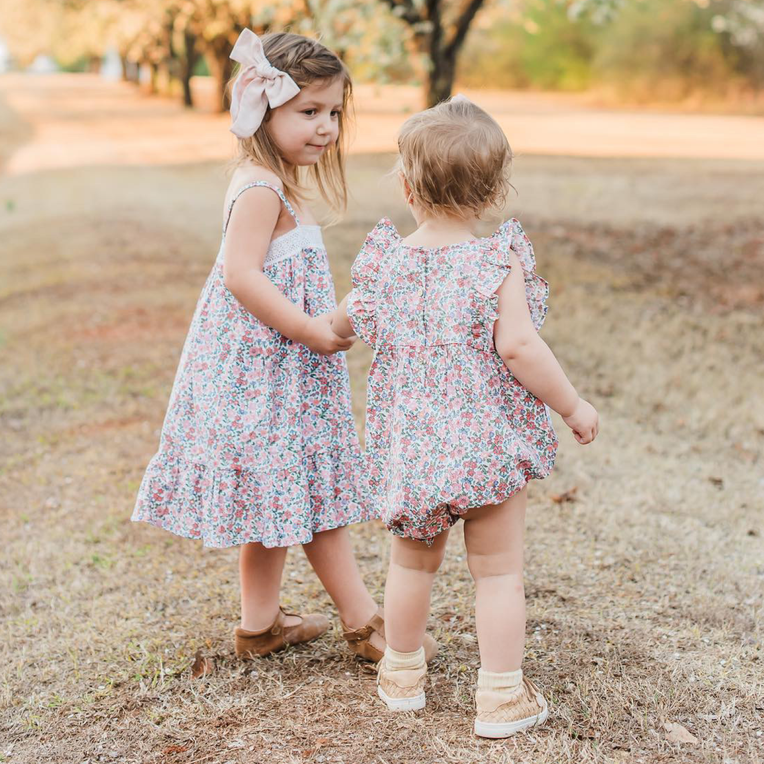 Two young children wearing floral outfits stand outdoors on a grassy path. The older child wears a dress and bow, holding hands with a younger child in a romper. Trees and warm sunlight fill the background.
