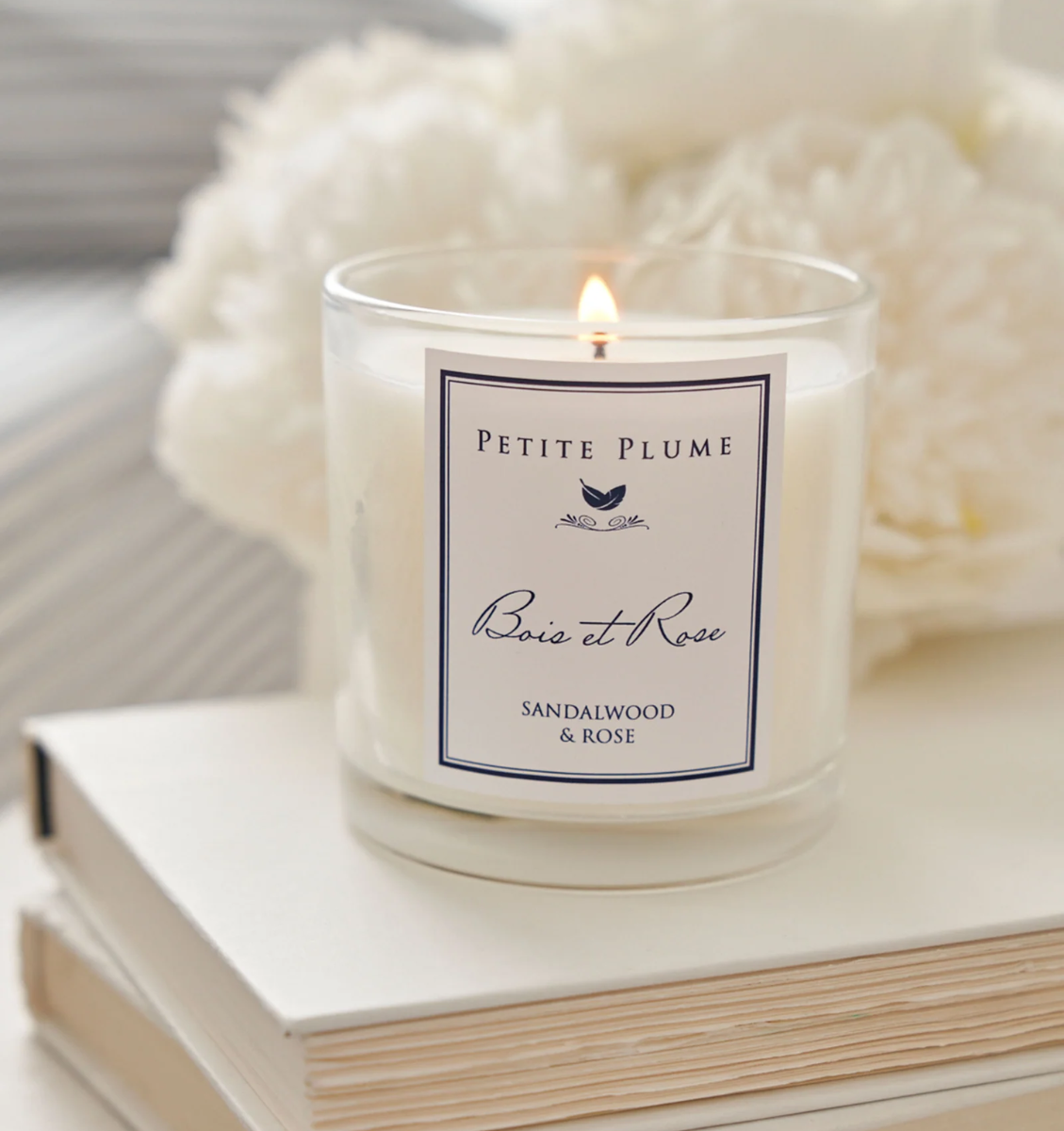 A lit white candle labeled Petite Plume Bois et Rose Sandalwood & Rose sits on a closed book, with white flowers softly blurred in the background.