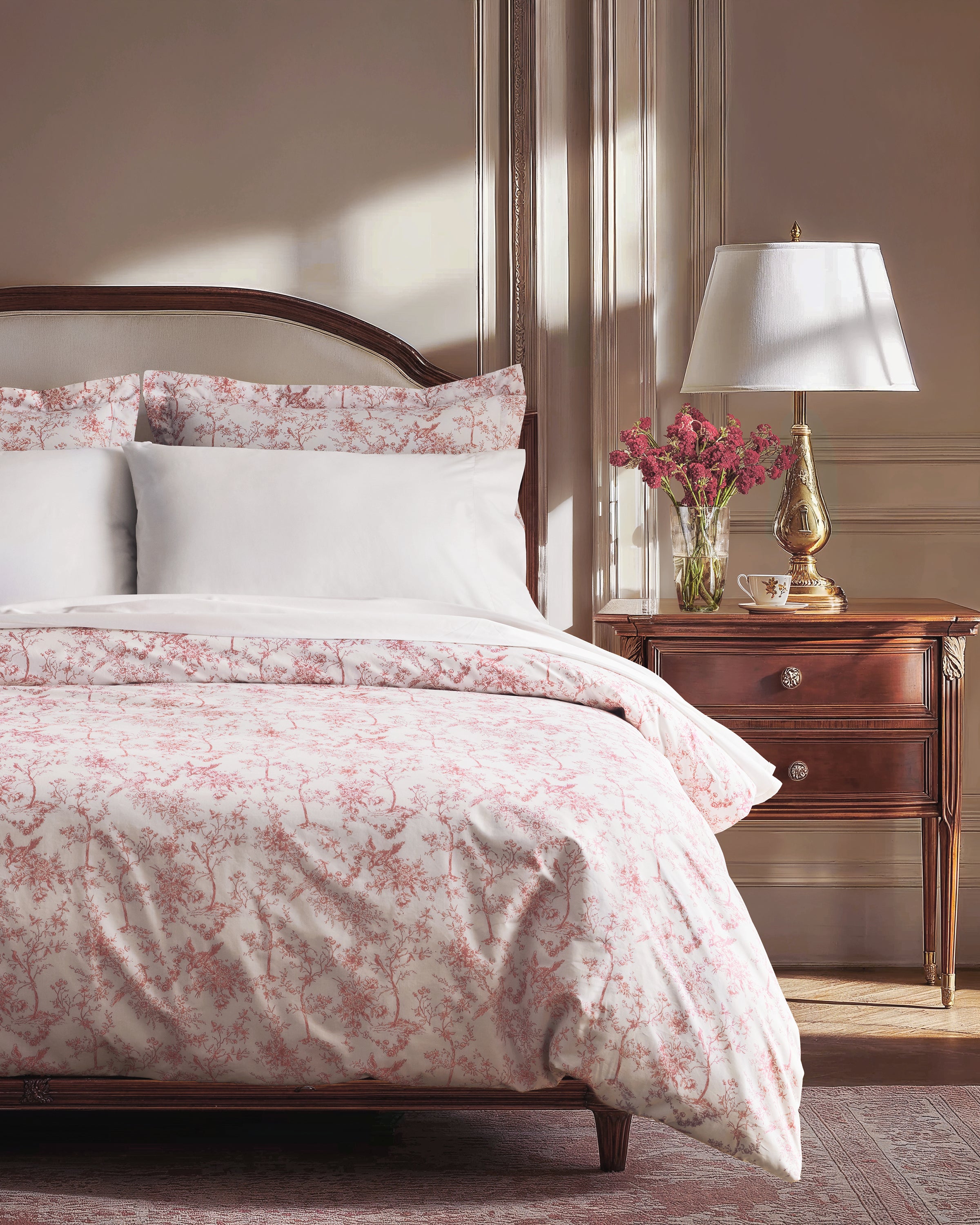 A neatly made bed features the Pink Timeless Toile Bedding Set with white sheets, a pink floral comforter, and two matching pillows against a curved, upholstered headboard in an elegant, classic bedroom.