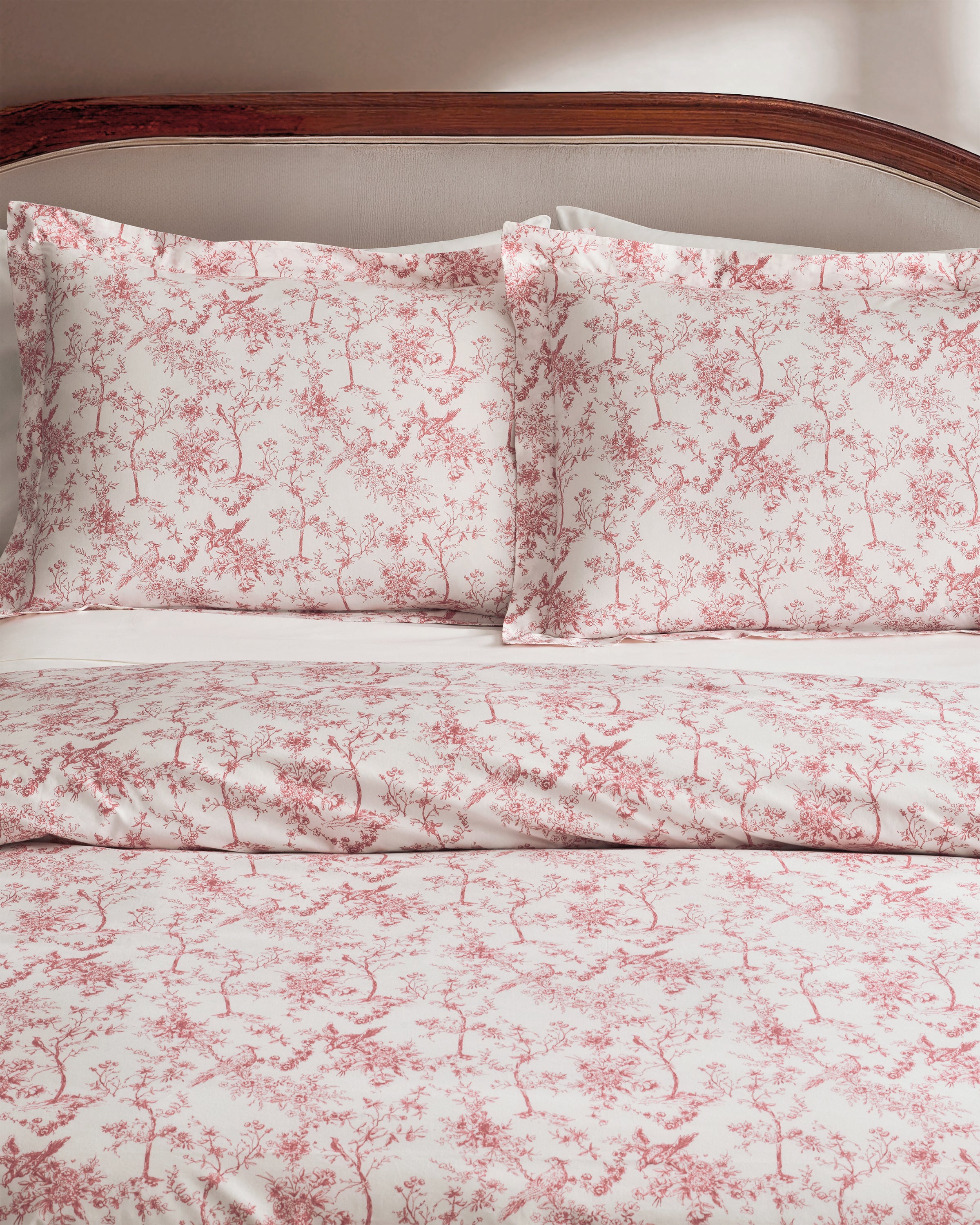 A neatly made bed features the Pink Timeless Toile Bedding Set with white sheets, a pink floral comforter, and two matching pillows against a curved, upholstered headboard in an elegant, classic bedroom.
