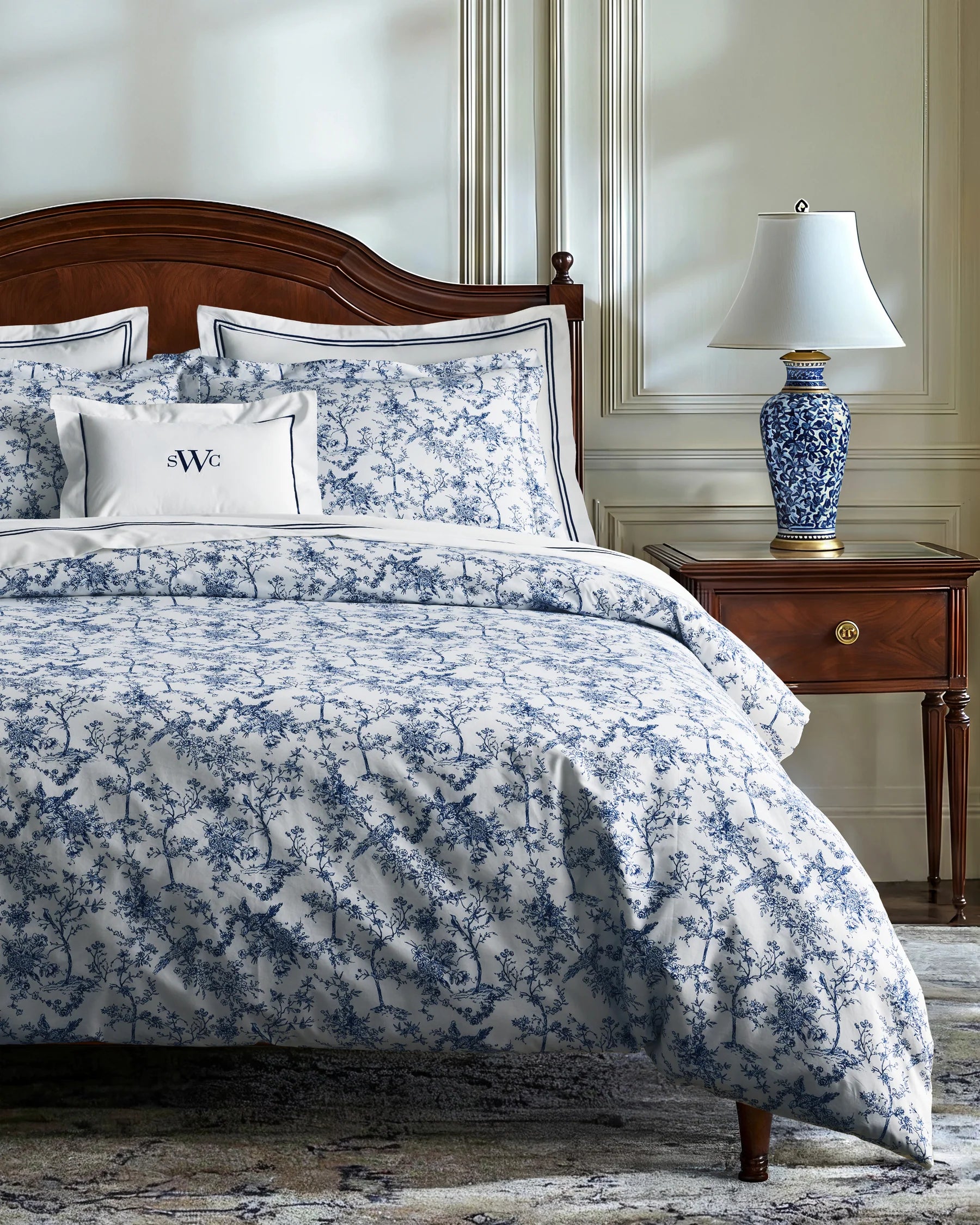 A neatly made bed features the Timeless Toile Bedding Set in white and blue with matching pillowcases, paired with a wooden headboard and a lamp on the nightstand in an elegant, well-lit bedroom.