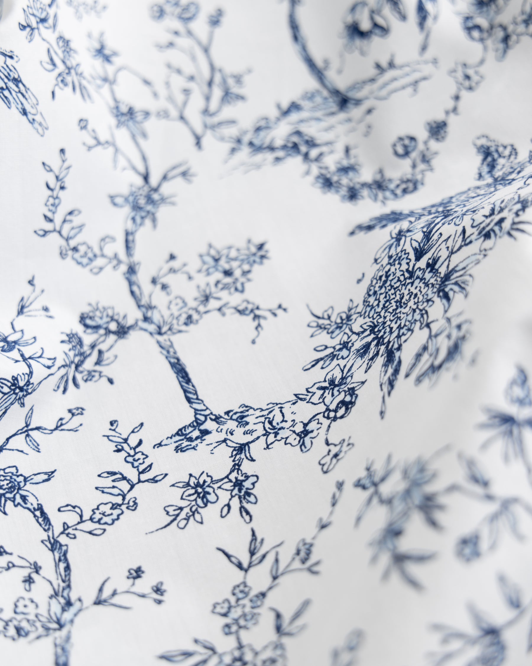 A close-up of the Luxe Premium Percale Duvet Set in Timeless Toile by Petite Plume shows an intricate blue floral pattern with branches, blossoms, and leaves, creating a detailed botanical motif on the soft, 100% cotton fabric.