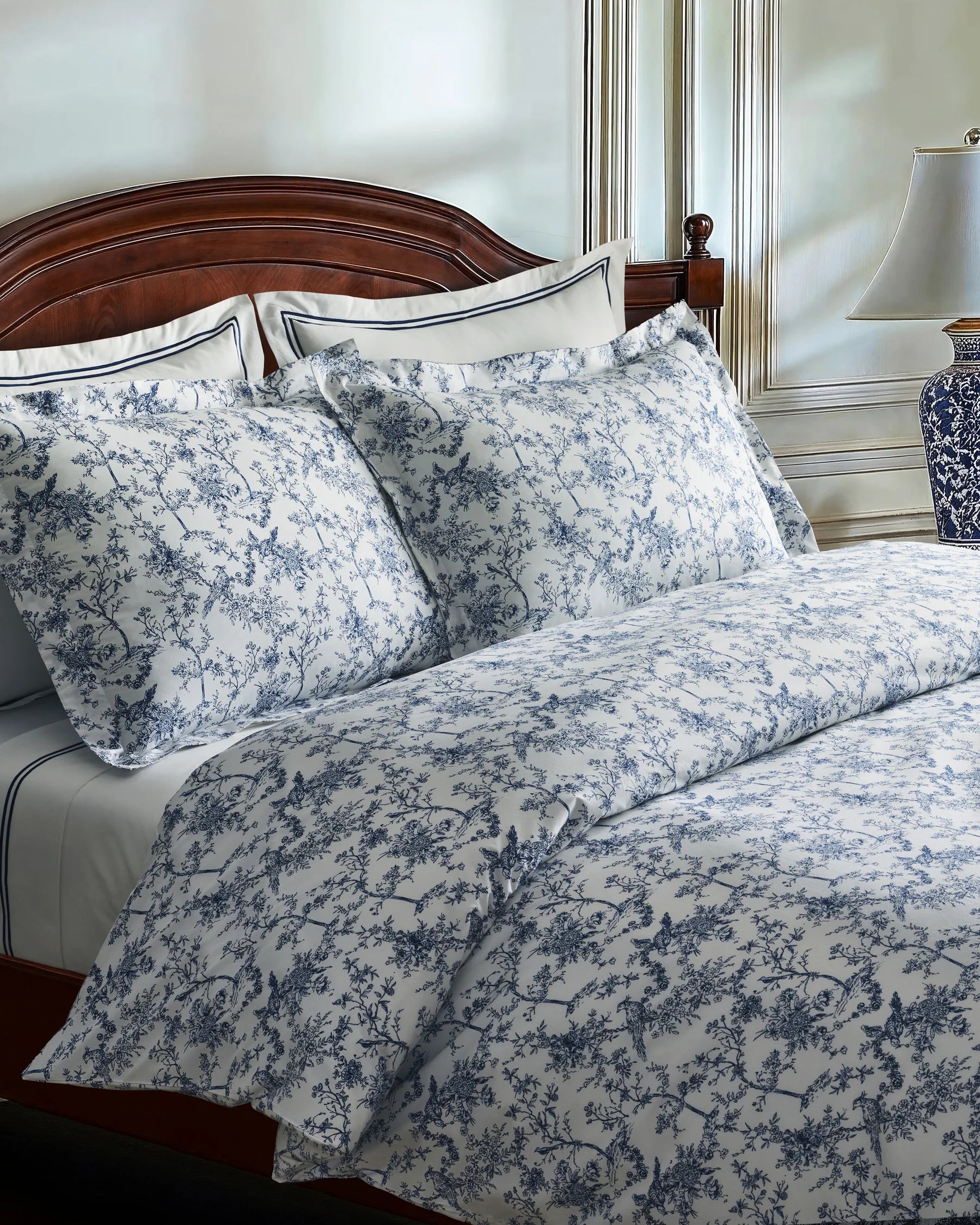A neatly made bed features the Timeless Toile Bedding Set in white and blue with matching pillowcases, paired with a wooden headboard and a lamp on the nightstand in an elegant, well-lit bedroom.