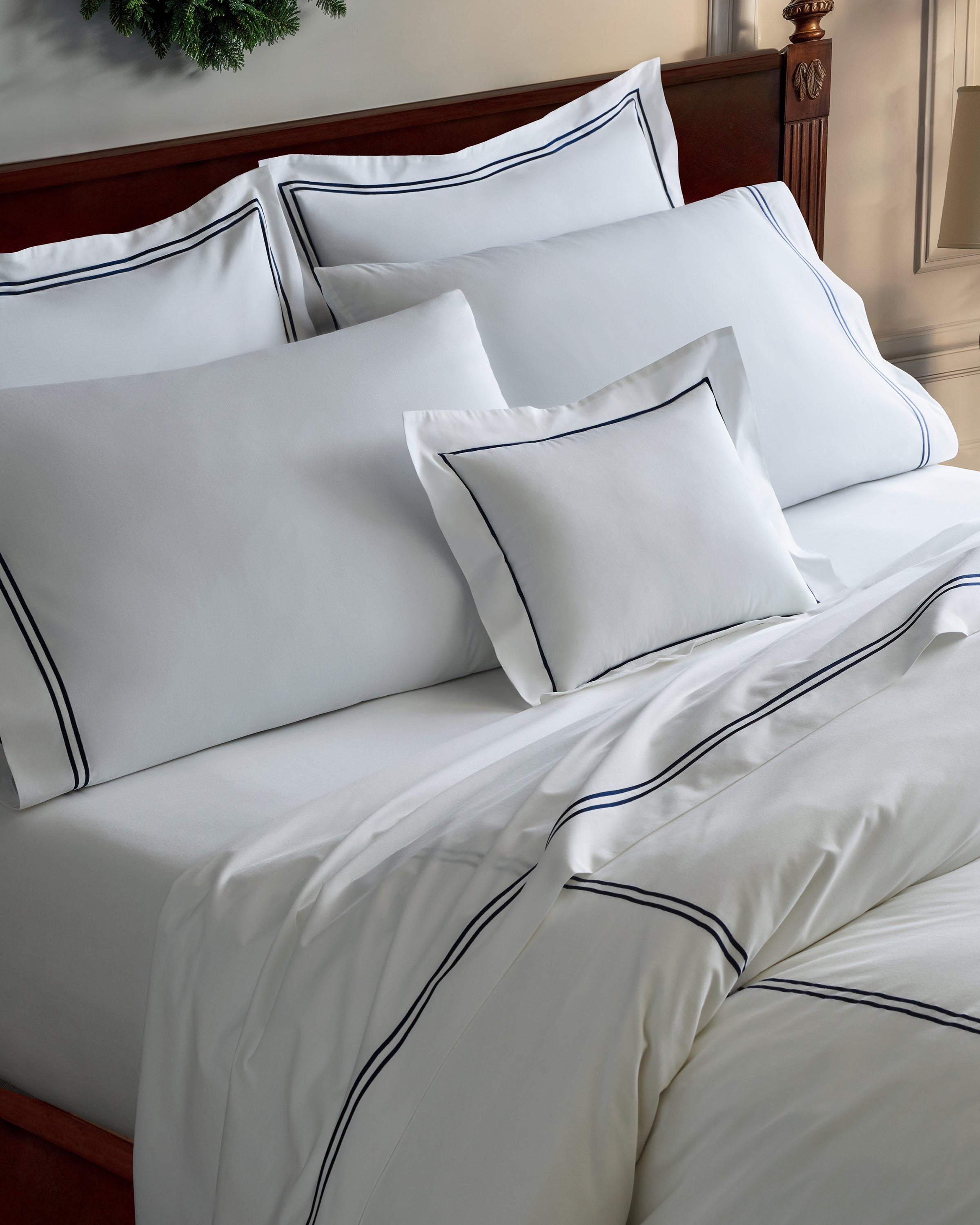 A neatly made bed with white bedding that features a dark, double-line border. The bed has multiple pillows and a comforter with matching trim, and the headboard is wooden.
