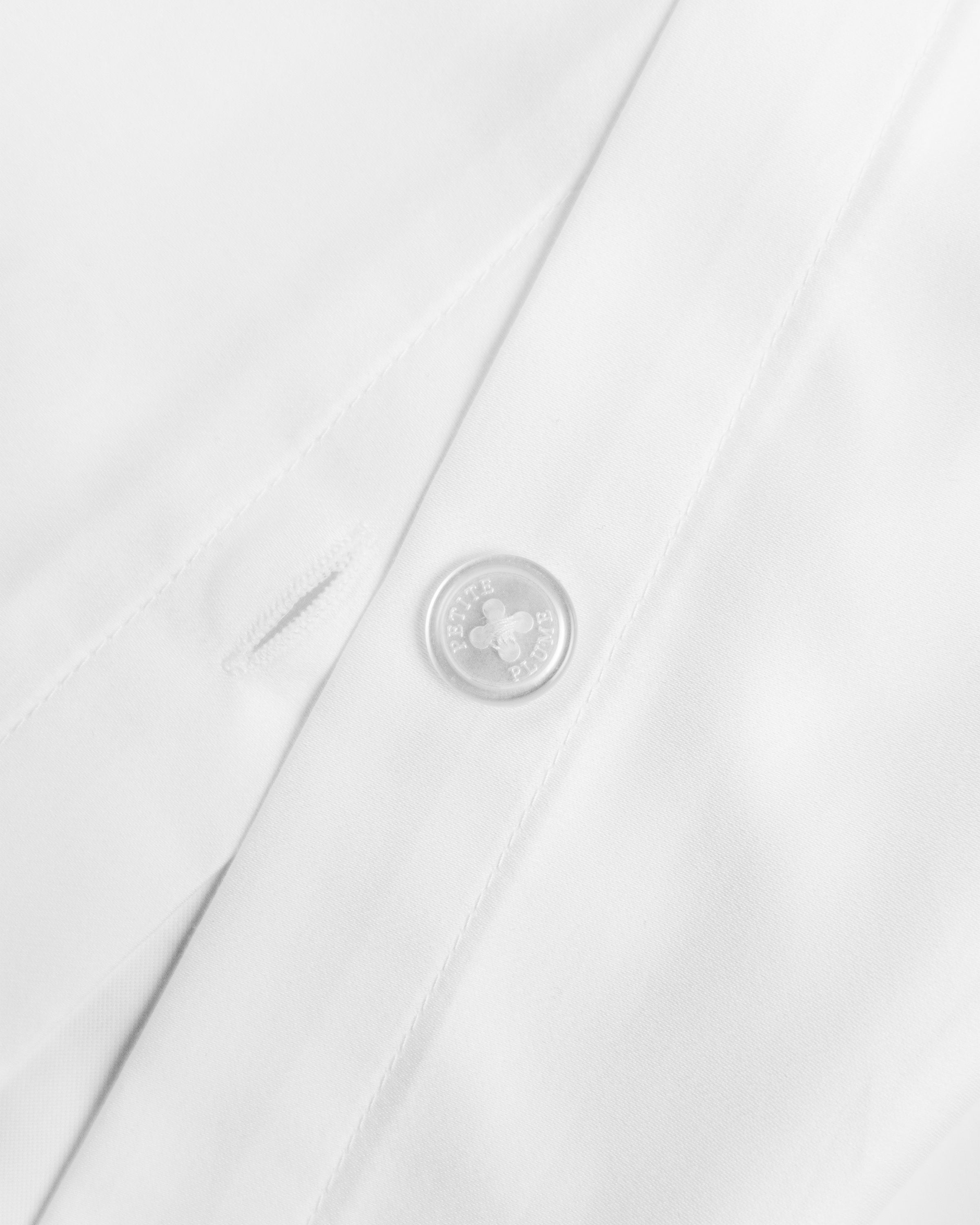Close-up of the Petite Plume Luxe Premium Percale Duvet Set in White, showing a button and buttonhole. The smooth, neatly stitched fabric resembles luxury sheets, radiating a clean and crisp texture. The button is circular with four holes.