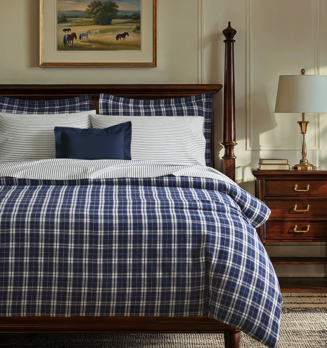 A neatly made bed with blue and white plaid bedding, matching striped pillows, and a dark wood headboard. A nightstand with a lamp and a framed painting of horses hang on the wall above the bed.