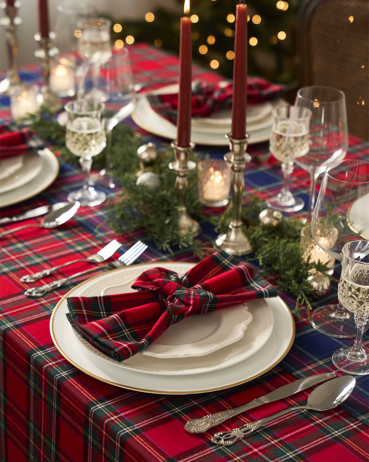 A festive tablescape features white plates on a Petite Plume Signature Twill Tablecloth in Imperial Tartan, accented with plaid napkins. Silver cutlery and crystal glasses complete the setting, while red candles and greenery add warmth beneath a softly lit Christmas tree.