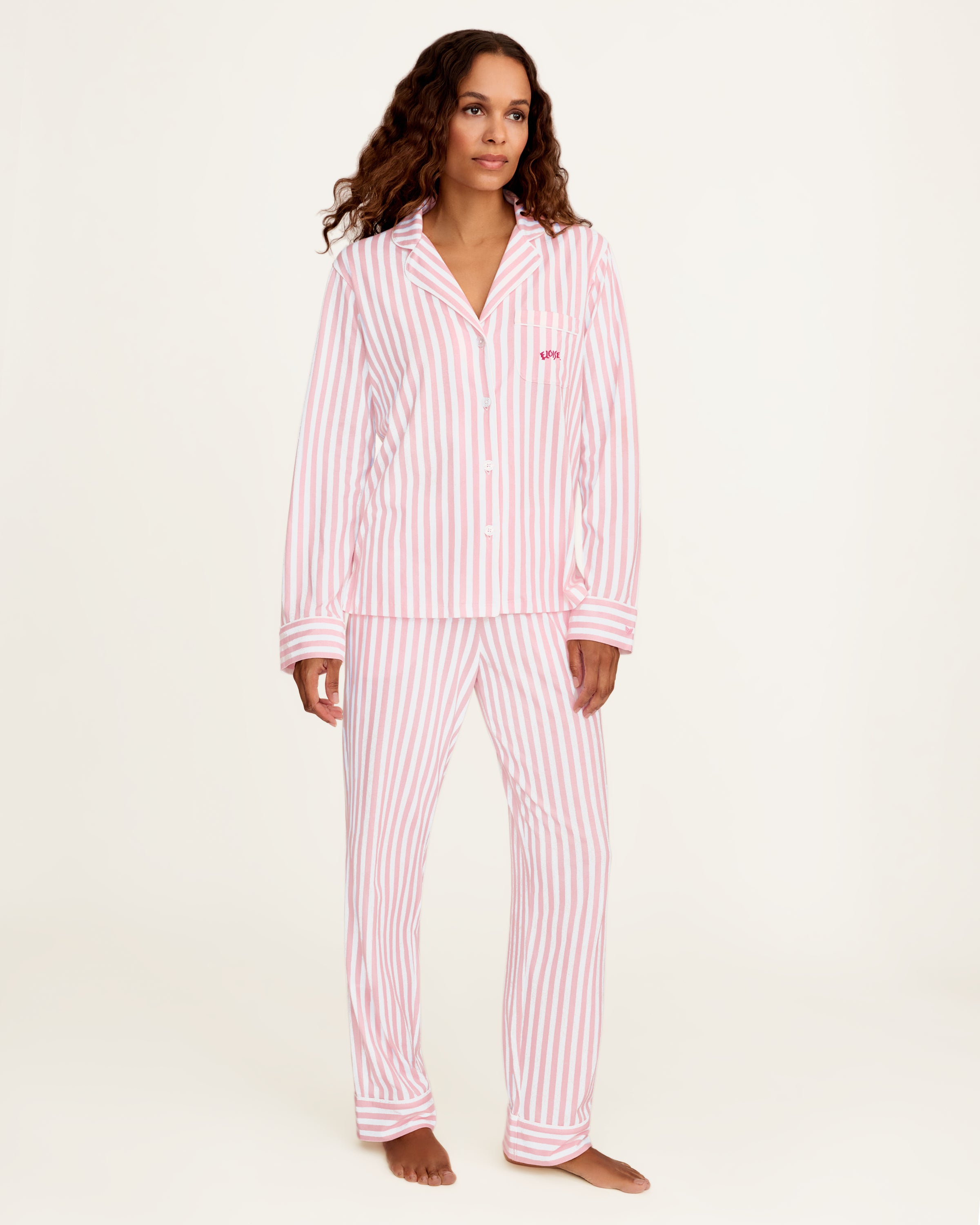 The Petite Plume x Eloise Women's Pima Pajama Set in Eloise Classic Stripe features pink and white vertical stripes, a long-sleeve button-up top with a collar, chest pocket with red embroidery, and matching pants.