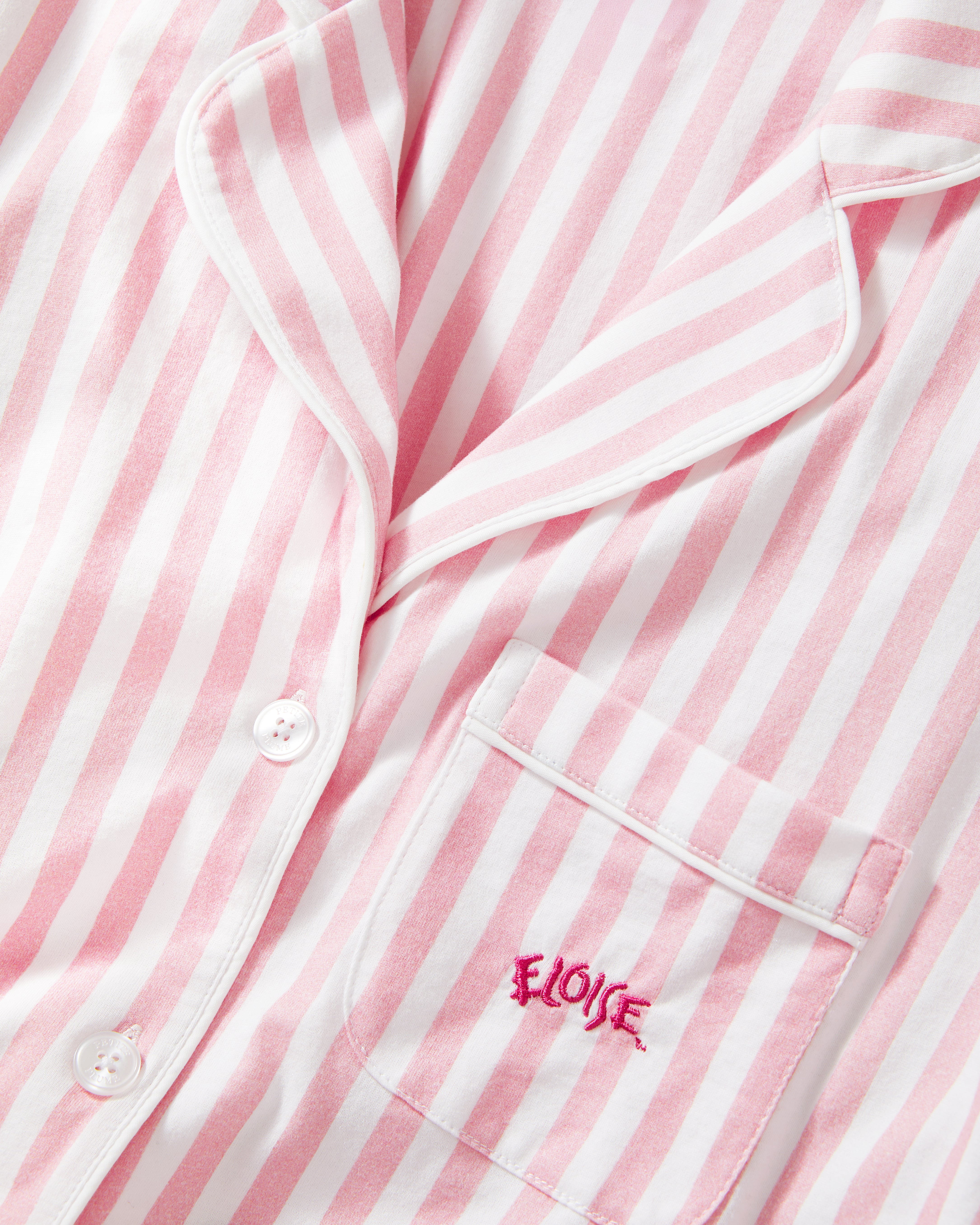 The Petite Plume x Eloise Women's Pima Pajama Set in Eloise Classic Stripe features pink and white vertical stripes, a long-sleeve button-up top with a collar, chest pocket with red embroidery, and matching pants.