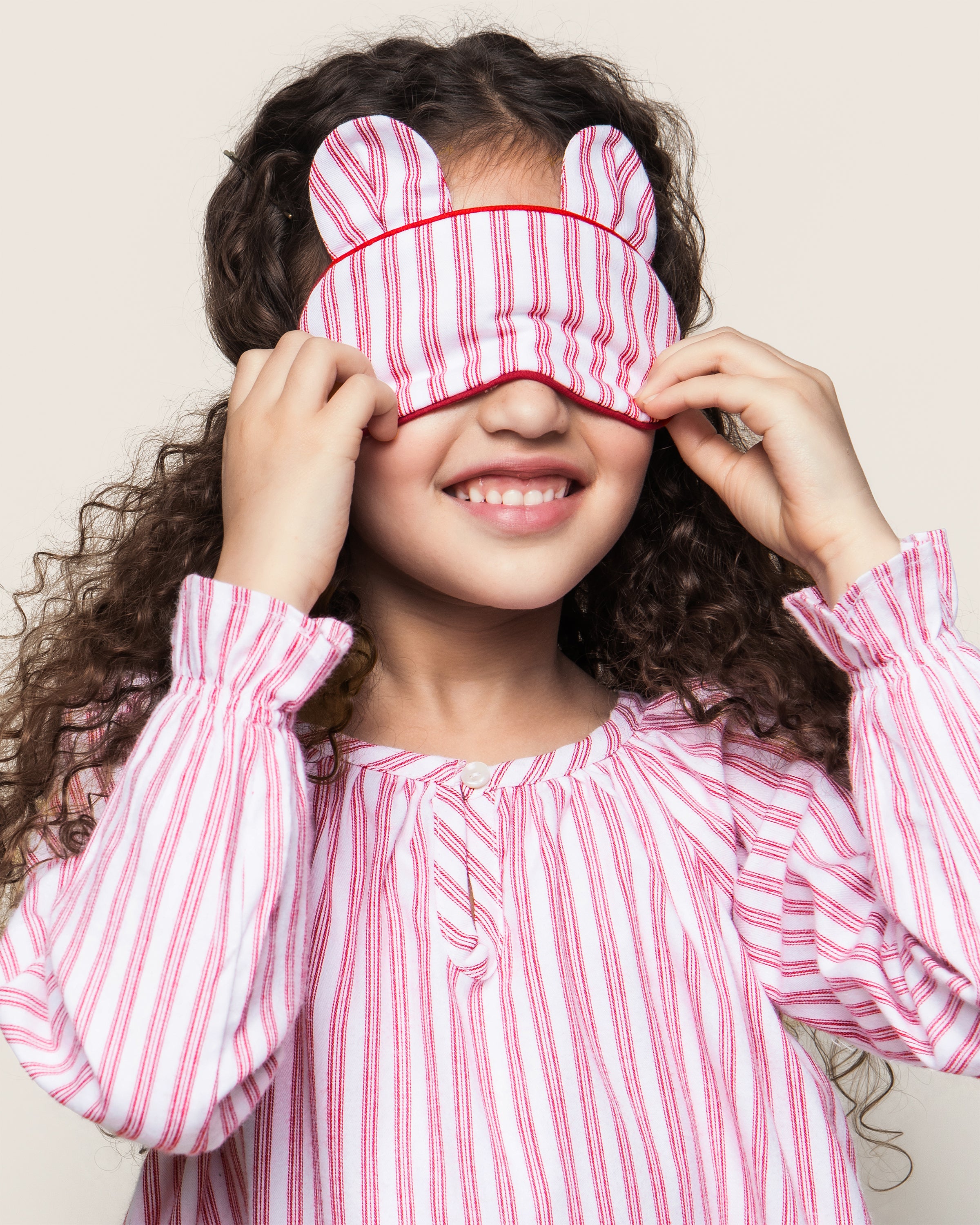 A young girl with curly hair smiles as she adjusts the Kids Bear-y Sleep Mask in Antique Red Ticking by Petite Plume, featuring bear ears. She sports a matching pink and white striped pajama top, adding charm to her luxurious sleepwear.