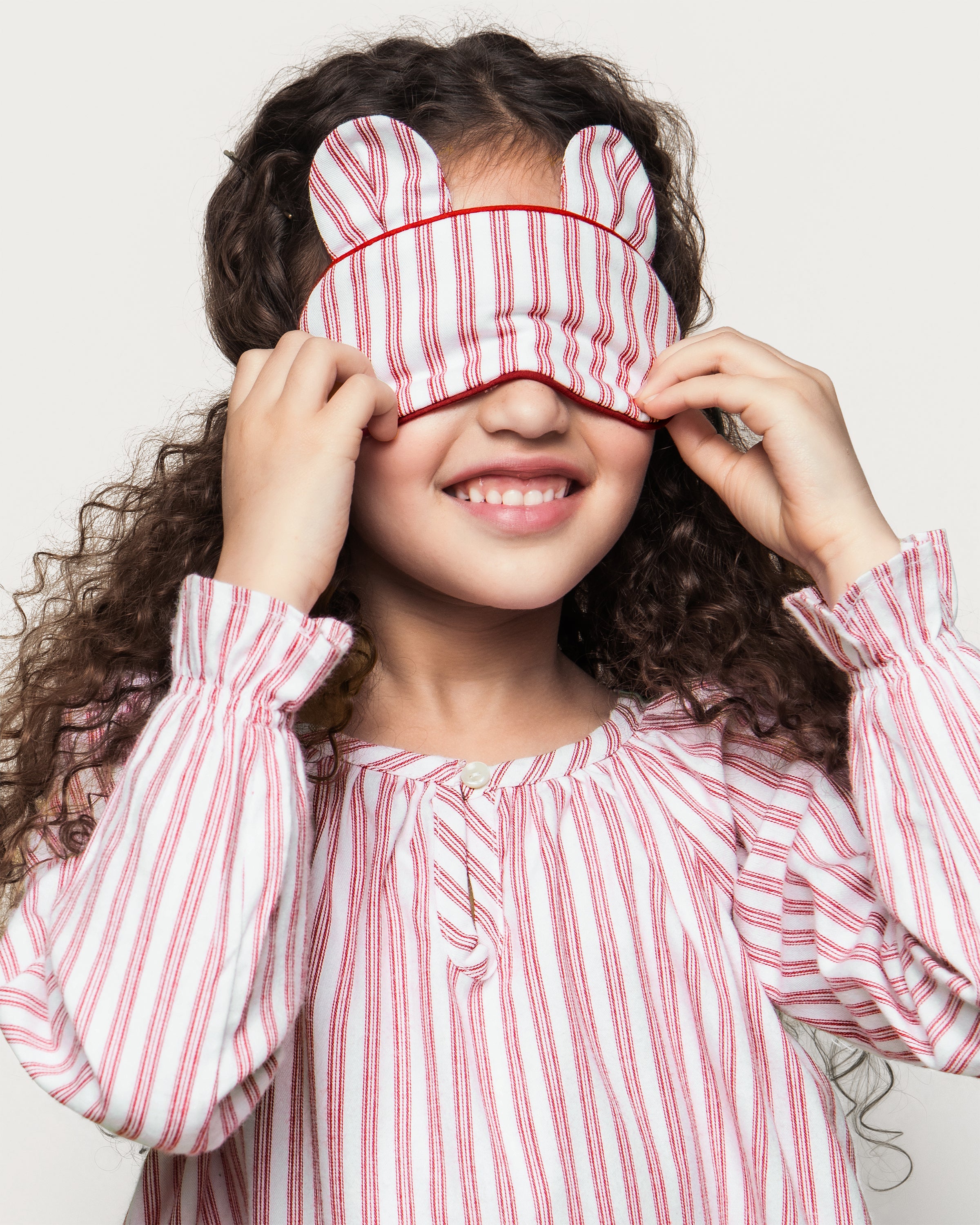 The Kid's Bear-y Sleep Mask in Antique Red Ticking features red and white stripes, two bear ear shapes on top, a white strap, and “PETITE” labeled on the side.