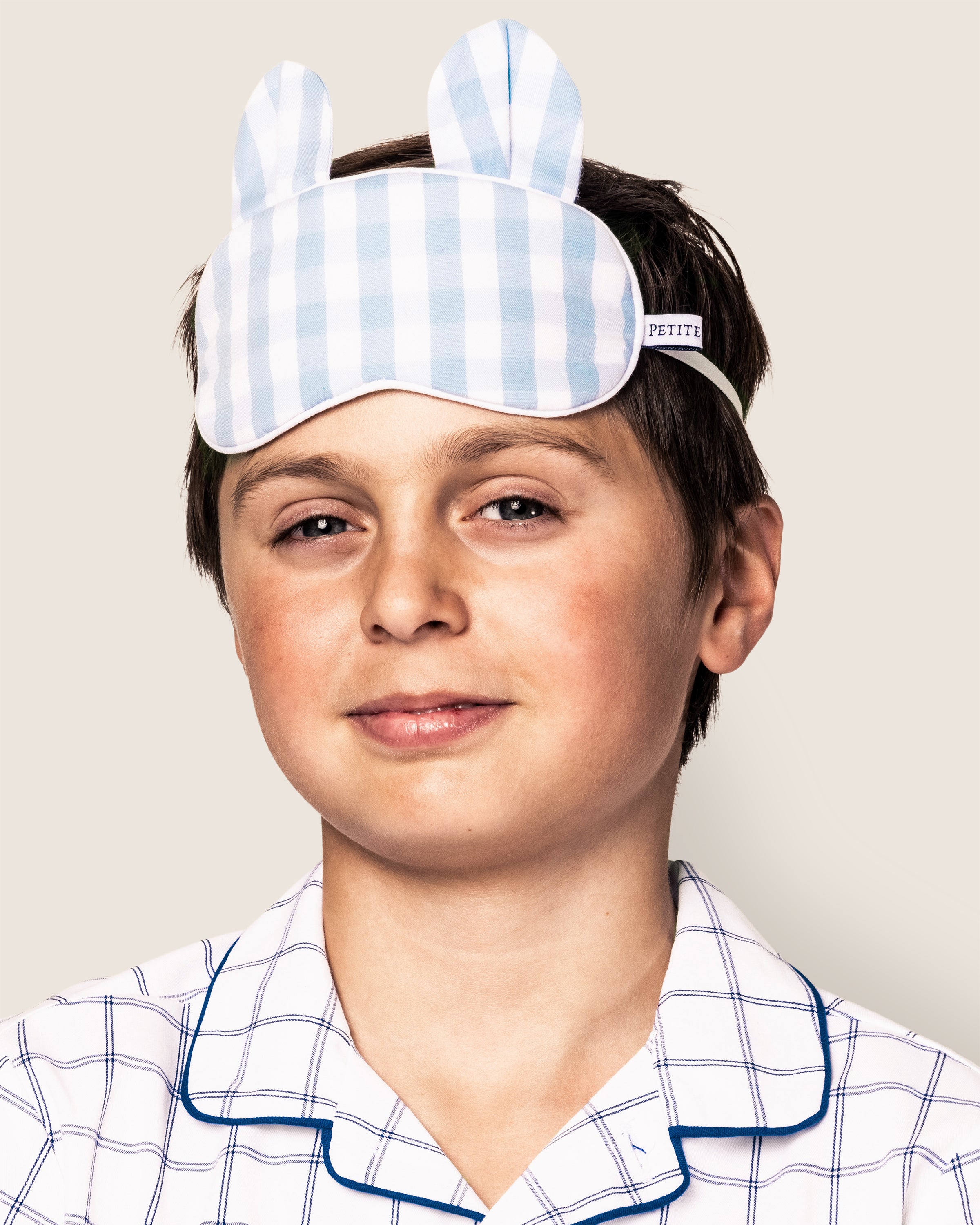 A child wearing a blue and white checkered pajama shirt and the Kids Twill Bear-y Sweet Sleep Mask in Light Blue Gingham with bunny ears from Petite Plume smiles at the camera against a plain light background.