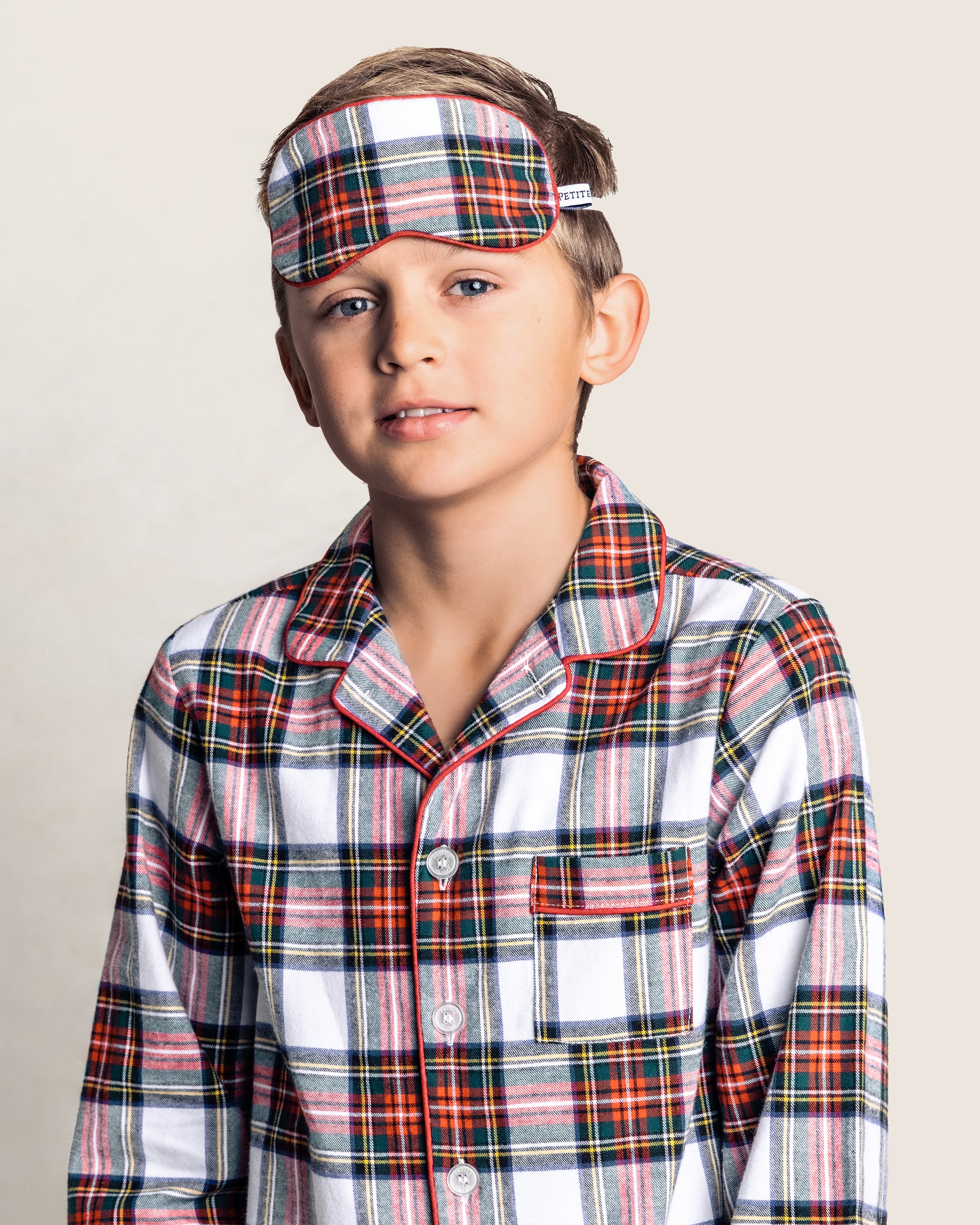 A young boy in Petite Plumes Kids Brushed Cotton Sleep Mask in Balmoral Tartan, alongside plaid pajamas, stands against a plain background. His short hair and neutral expression enhance the serene vibe of this perfectly coordinated ensemble.