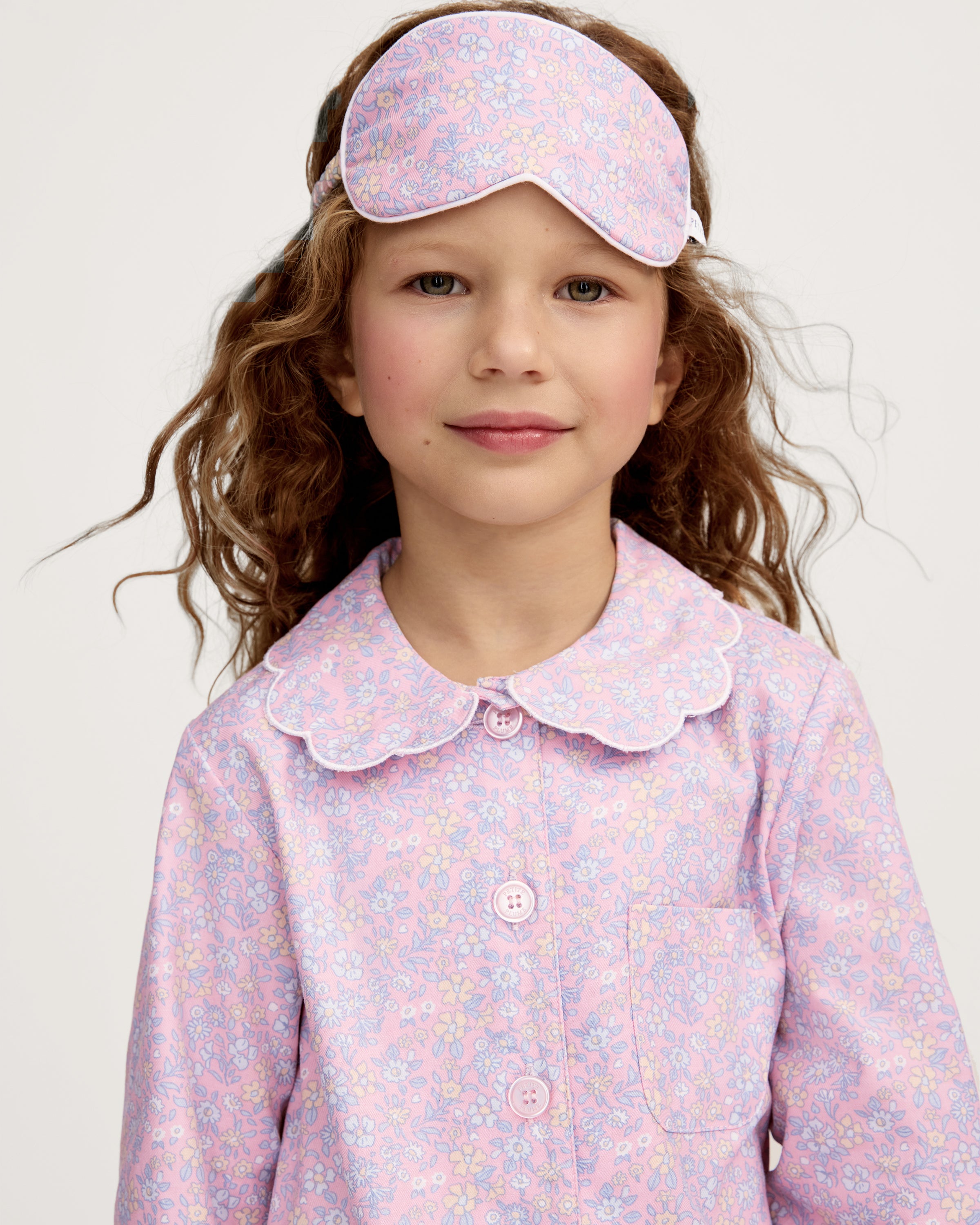 A young girl with wavy brown hair wears the Kid's Twill Sleep Mask in Fleur Blush on her forehead, paired with a matching pastel pink floral pajama set, standing against a plain light background.