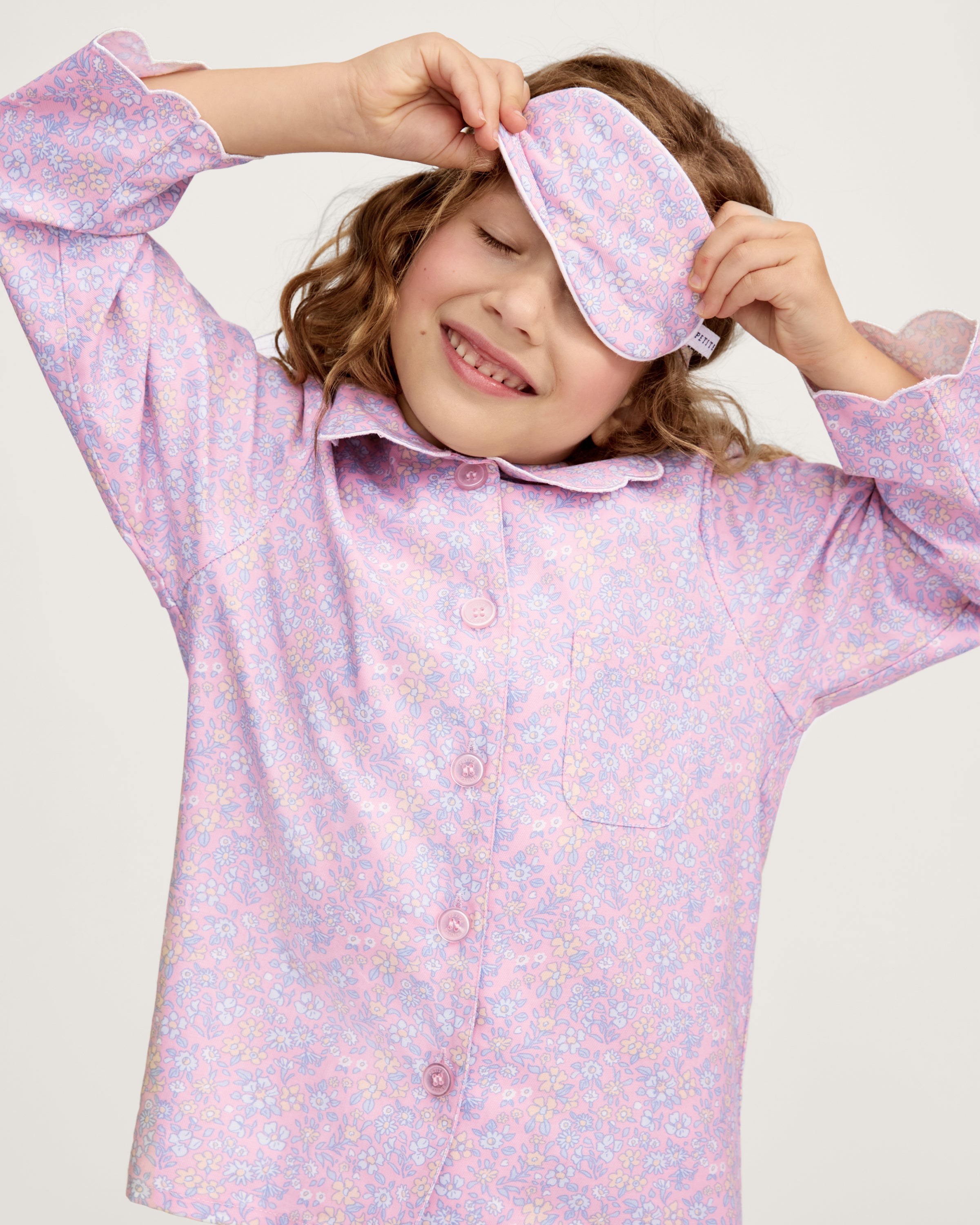 A young girl with wavy brown hair wears the Kid's Twill Sleep Mask in Fleur Blush on her forehead, paired with a matching pastel pink floral pajama set, standing against a plain light background.