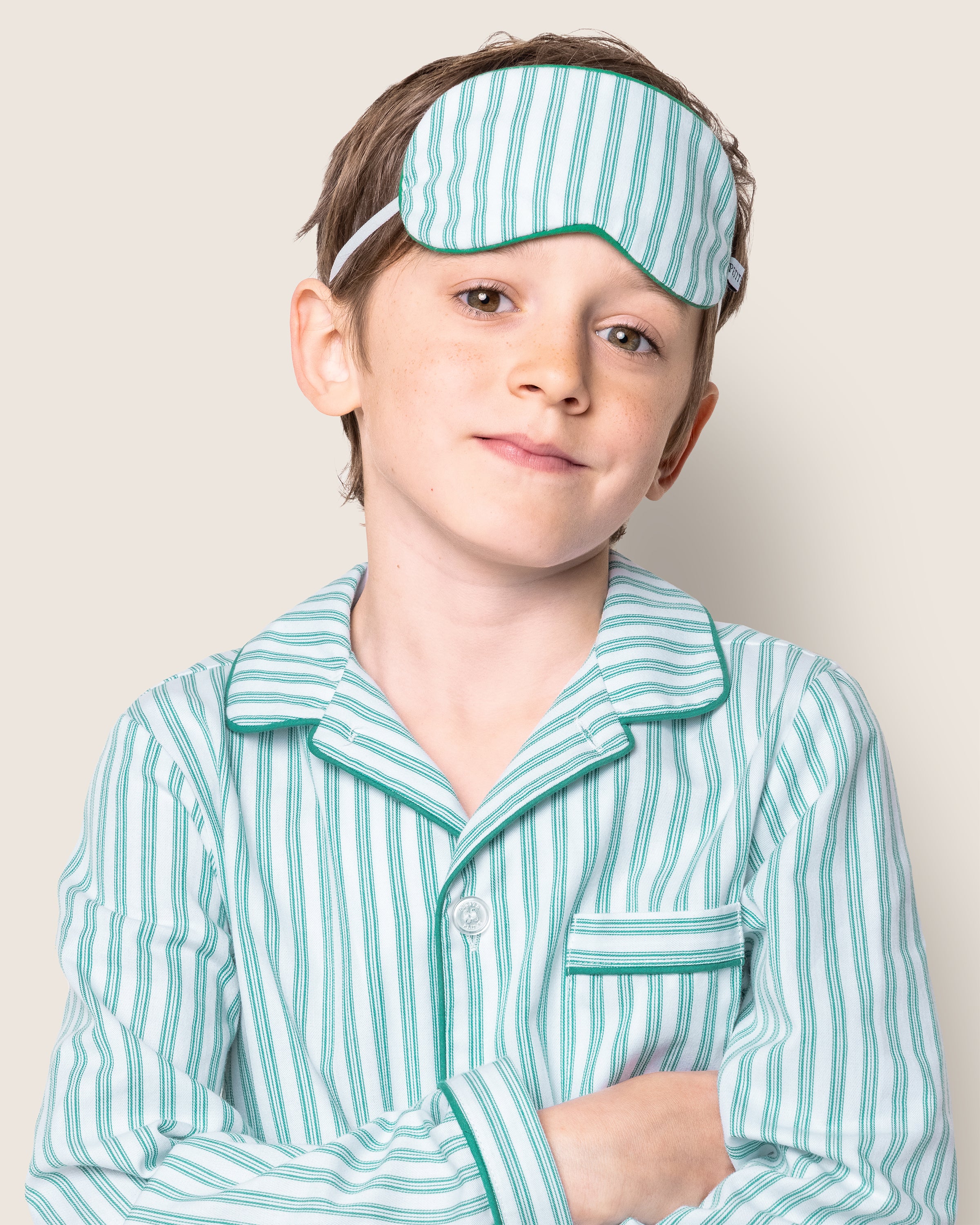 A young boy, wearing Petite Plumes luxurious green and white striped pajamas and the Kids Twill Sleep Mask in Emerald Ticking, stands with arms crossed and a slight smile against a light background.