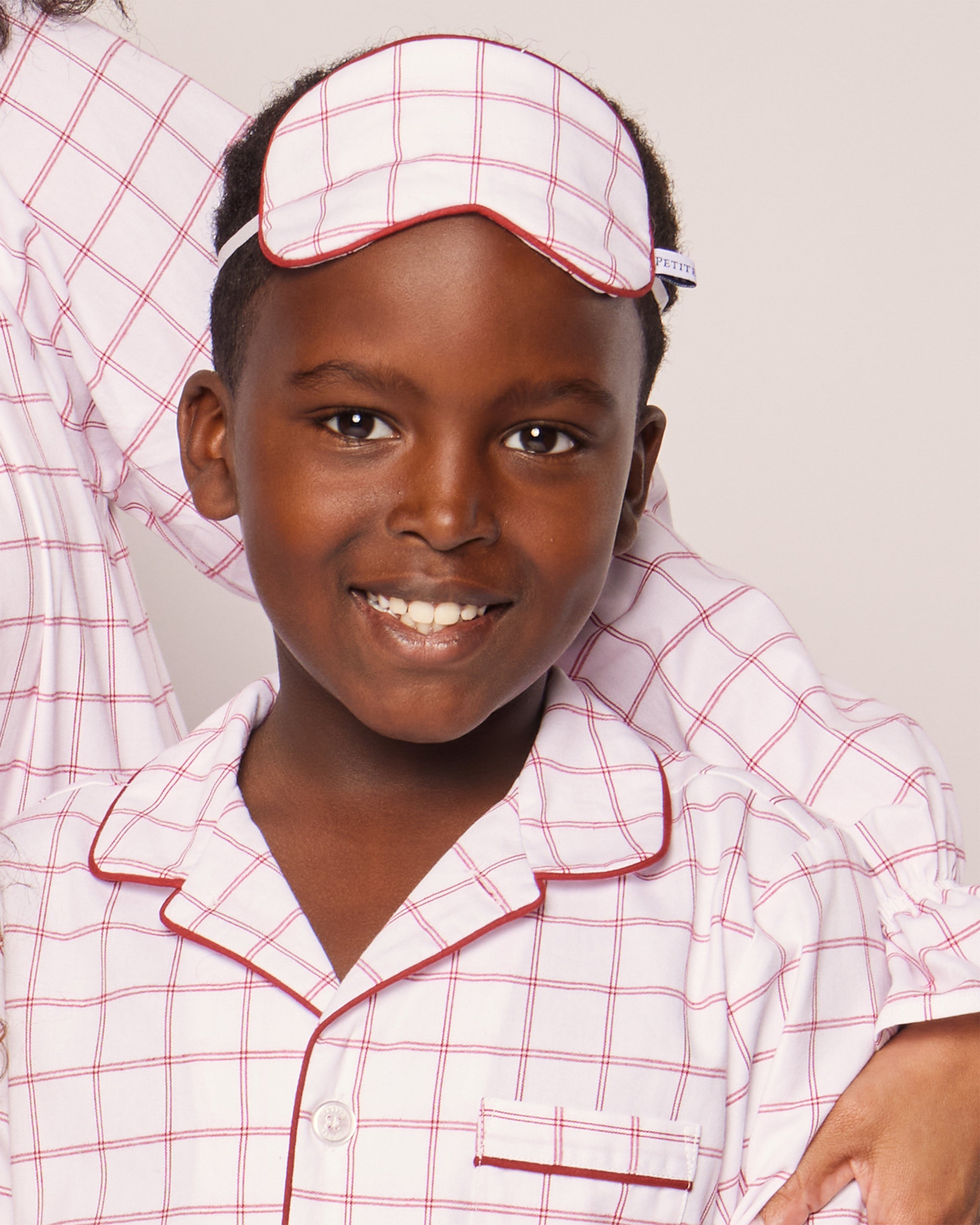 A smiling child wears Petite Plumes Kids Twill Sleep Mask in Garnet Tattersall, a luxurious white and red checkered pajama set with elegant red piping, a small breast pocket, and an arm draped over their shoulder.