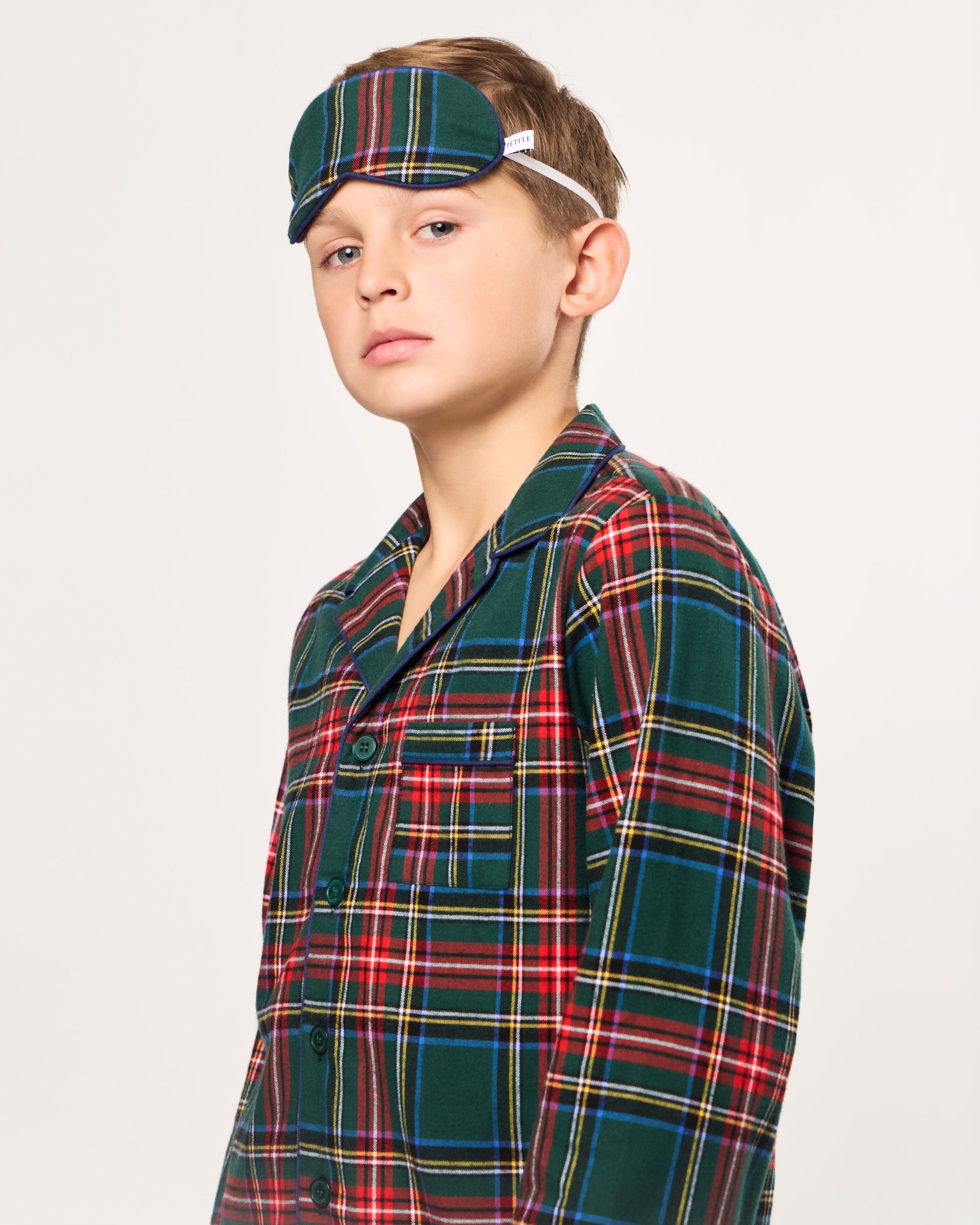 The Kid's Twill Sleep Mask in Kensington Tartan features red, green, yellow, blue, and white plaid, an elastic strap, and a small white PETITE tag on the right side.