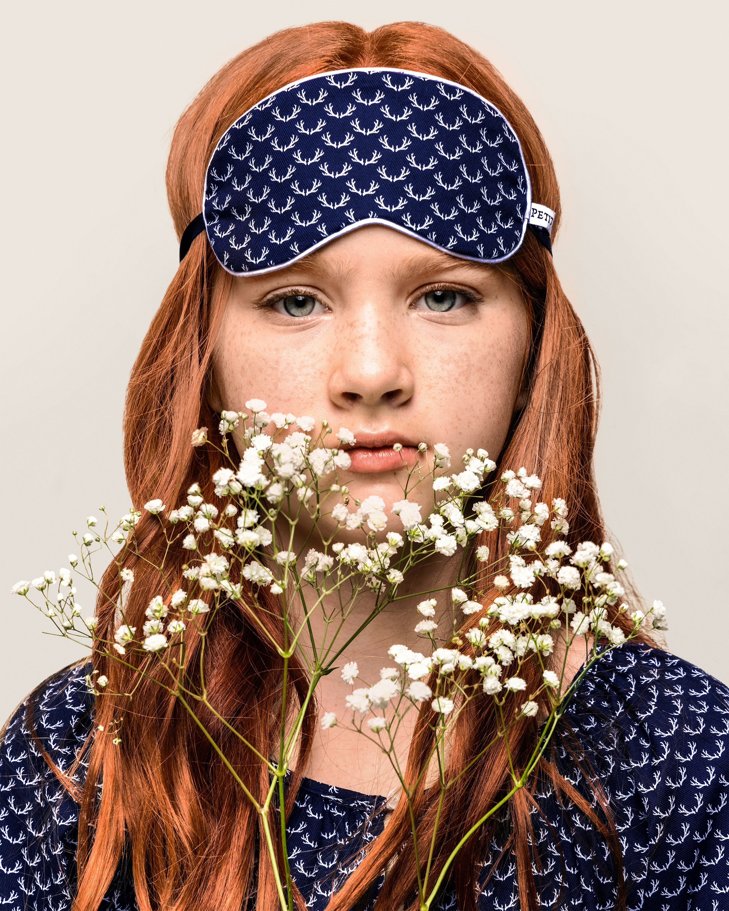 A person with long red hair wears luxurious sleepwear, featuring a navy blue mask from Petite Plume’s Kids Twill Sleep Mask in Nordic Antlers, matching their shirt. They stand against a neutral background, holding white babys breath, and the eye mask boasts elegant white patterns.