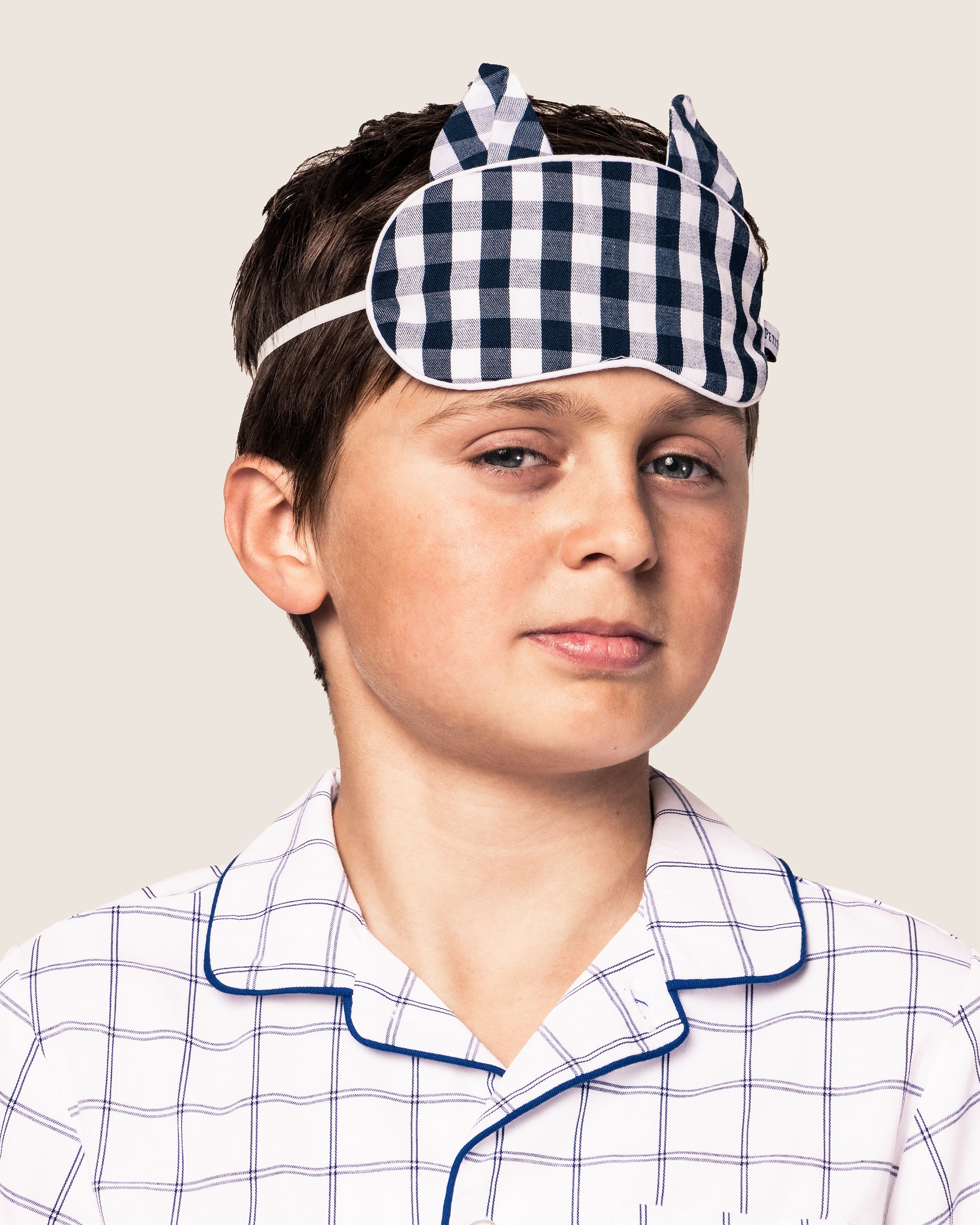 A young boy in Petite Plumes luxurious sleepwear, featuring the Kids Twill Kitty Sleep Mask in Navy Gingham with ear-like details and matching pajamas, looks directly at the camera with a slight smile. The background is a plain light gray.
