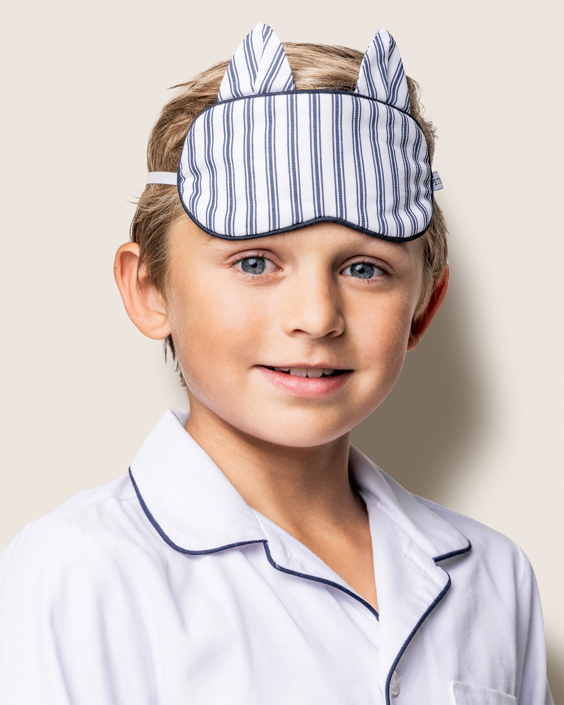 A boy in white pajamas with dark piping and a monogram beams with joy, donning the Petite Plume Kids Twill Kitty Sleep Mask in Navy French Ticking, set against a cream background—a perfect gift for imaginative little dreamers.