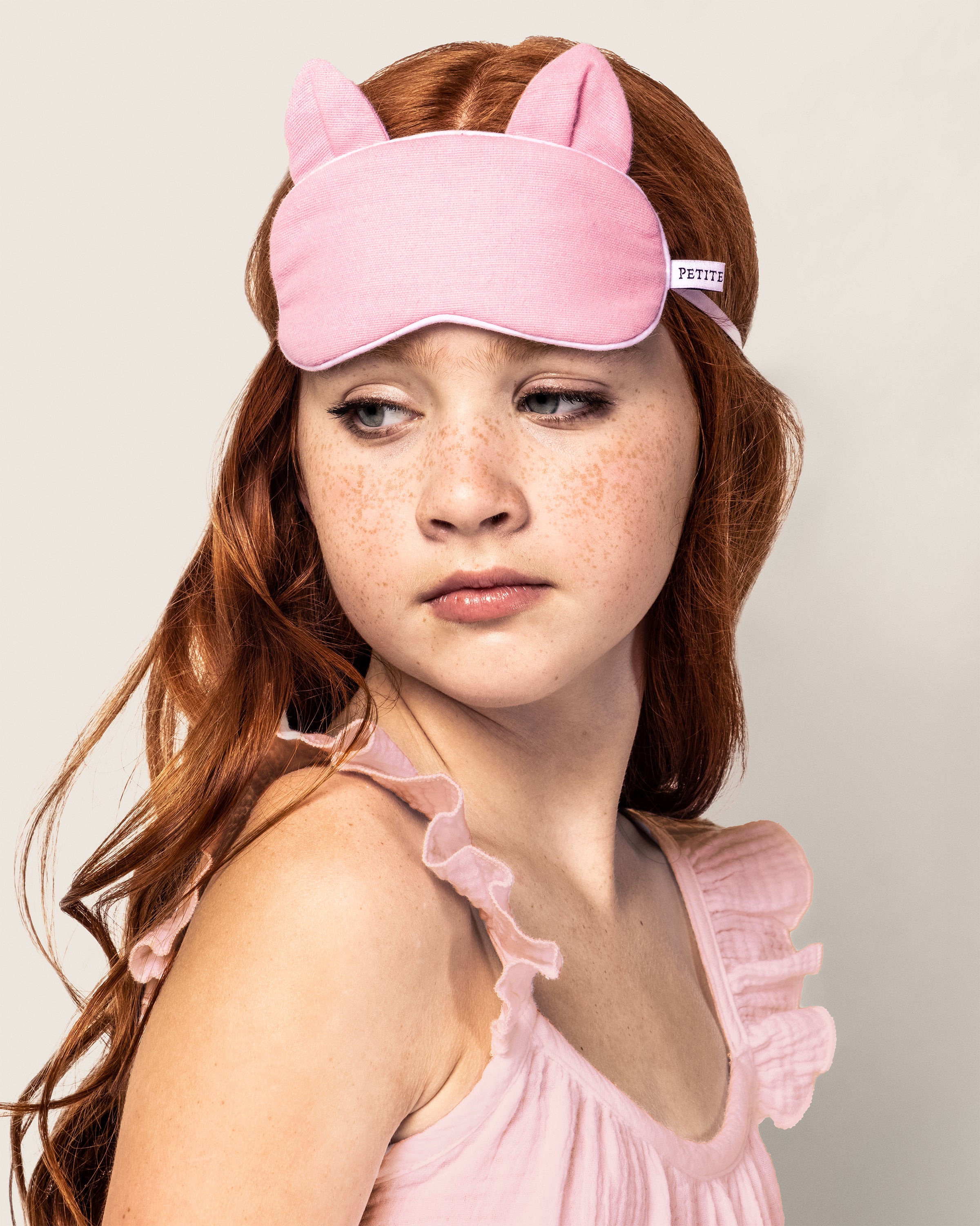 A young girl with long red hair wears the Kids Flannel Kitty Eye Mask in Pink by Petite Plume, complementing her luxurious sleepwear. Gazing to the side, shes dressed in a sleeveless ruffled pink top against a plain background.