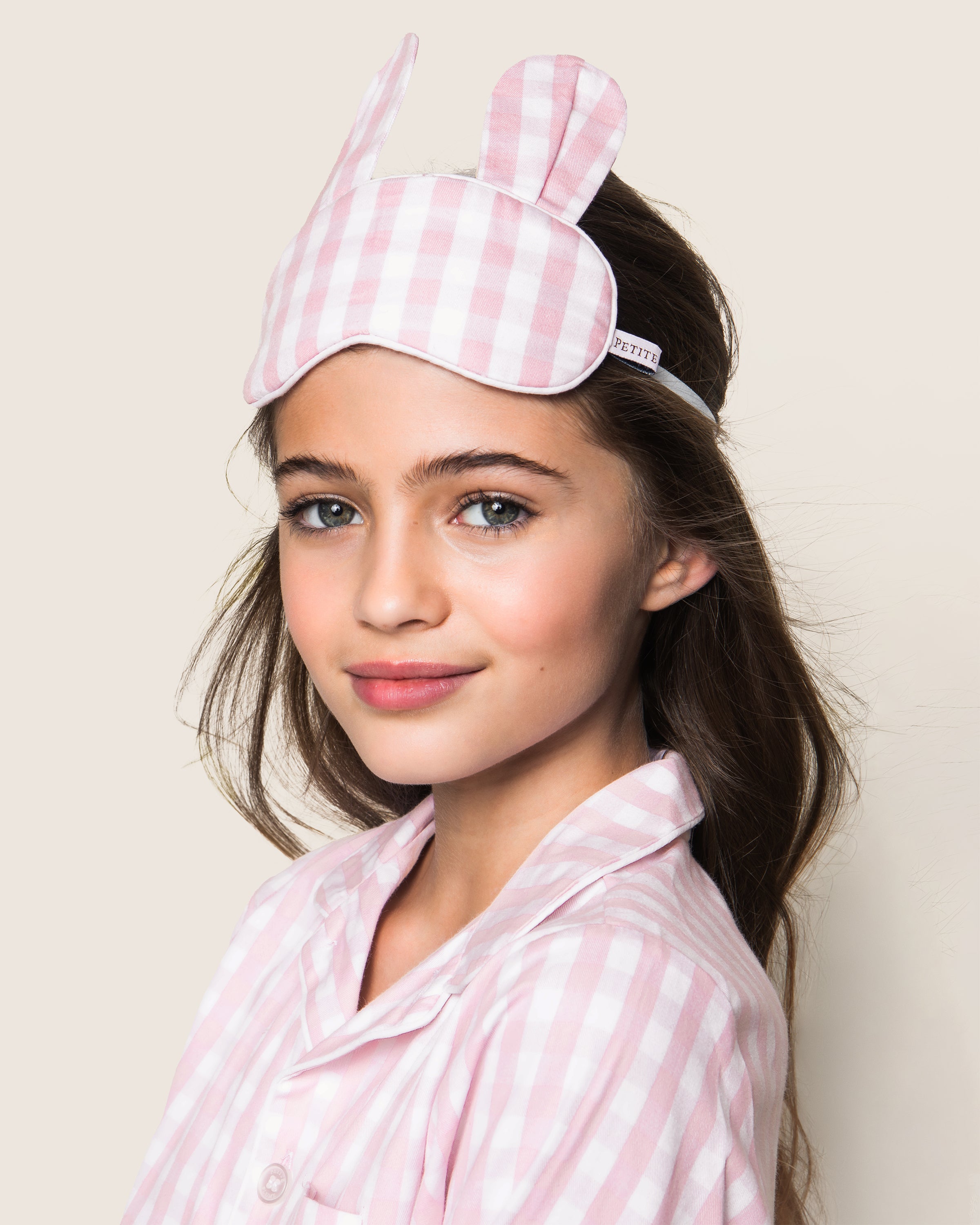 A girl smiles at the camera in her dreamland attire, wearing a pink and white checkered pajama set with a matching Petite Plume Kids Twill Bunny Sleep Mask in Pink Gingham. Her long dark hair cascades down against a plain, light background.