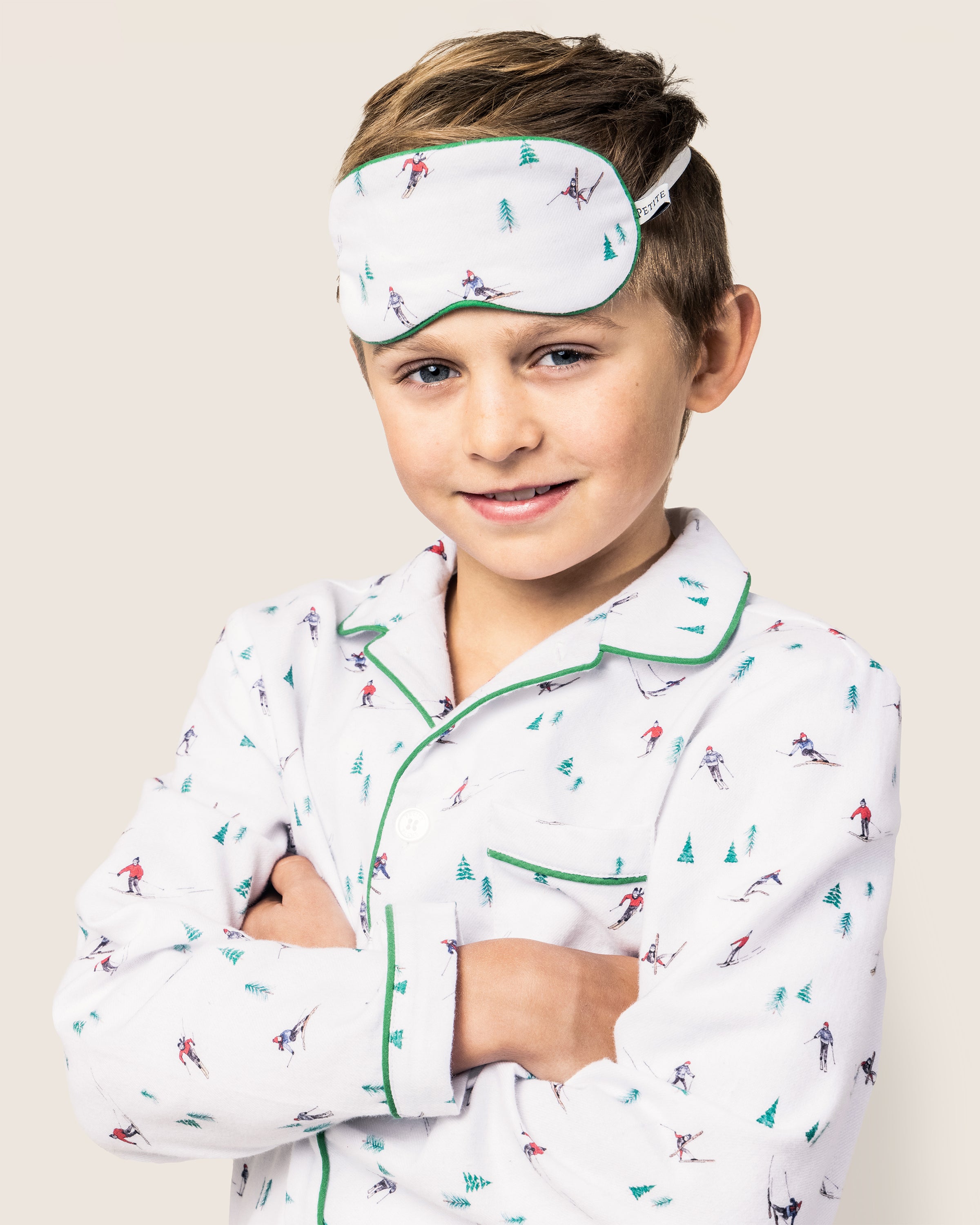 A child smiles gently in a luxurious Petite Plume white pajama set with colorful prints and green trim, arms crossed, as their Kids Flannel Sleep Mask in Après Ski rests on their forehead against a light background.