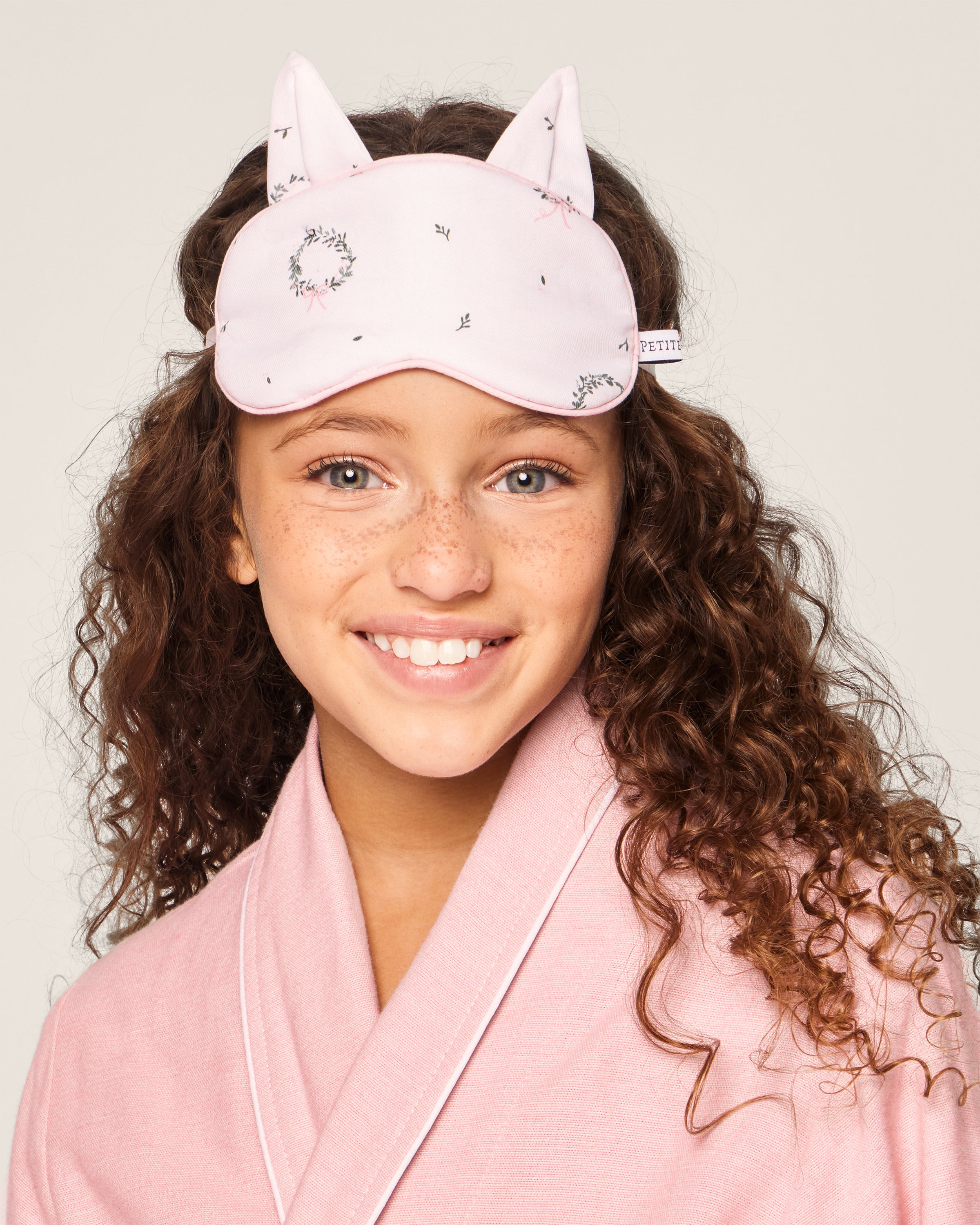 A smiling girl with curly hair wears Petite Plumes Kids Twill Kitty Eye Mask in Somerset Wreath. The neutral background highlights her joy and cozy look, evoking a luxurious dreamland feel.