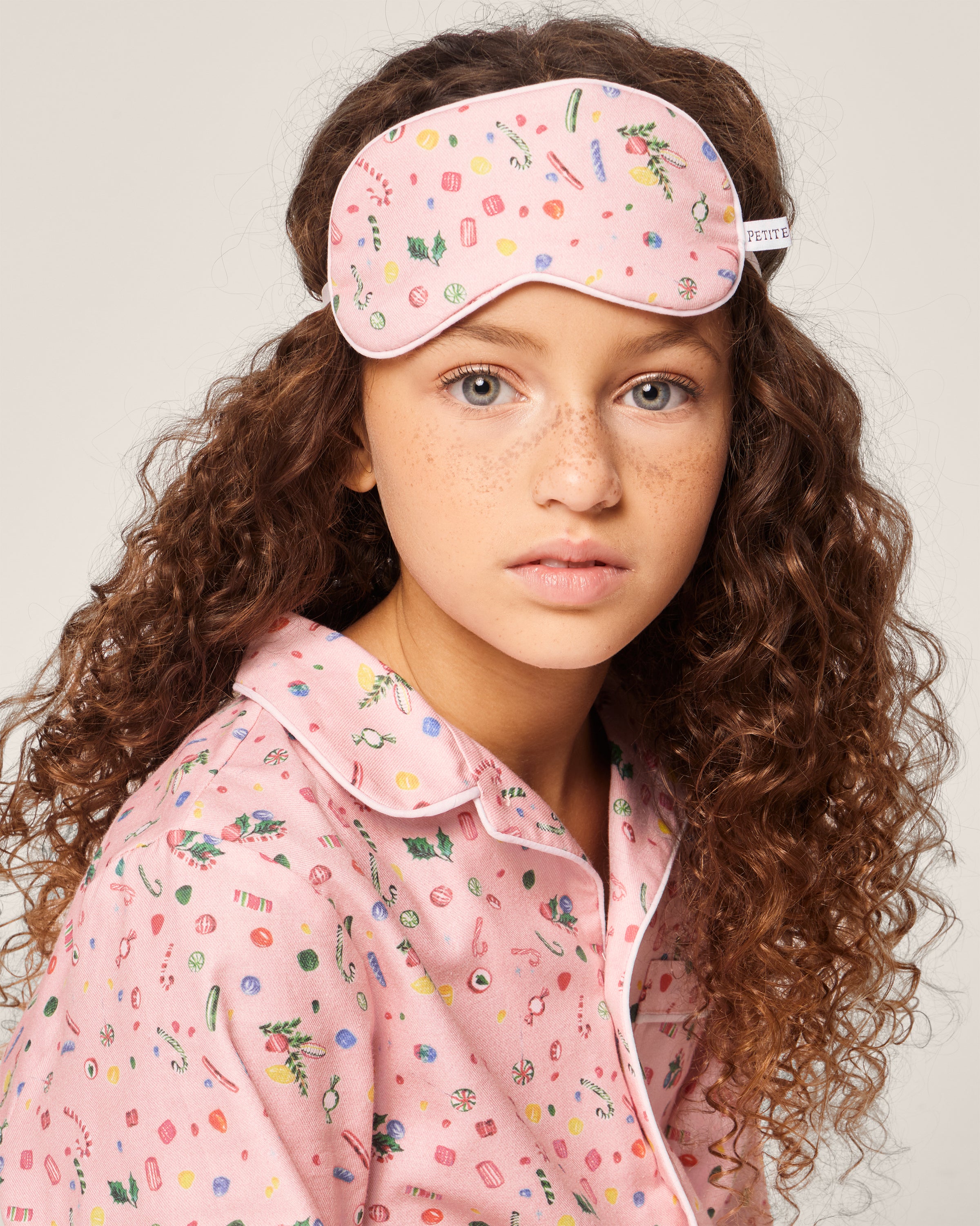 A young person with curly hair wears a Petite Plume Kids Twill Eye Mask in Vintage Sweets and matching colorful pajamas, set against a neutral gray background.