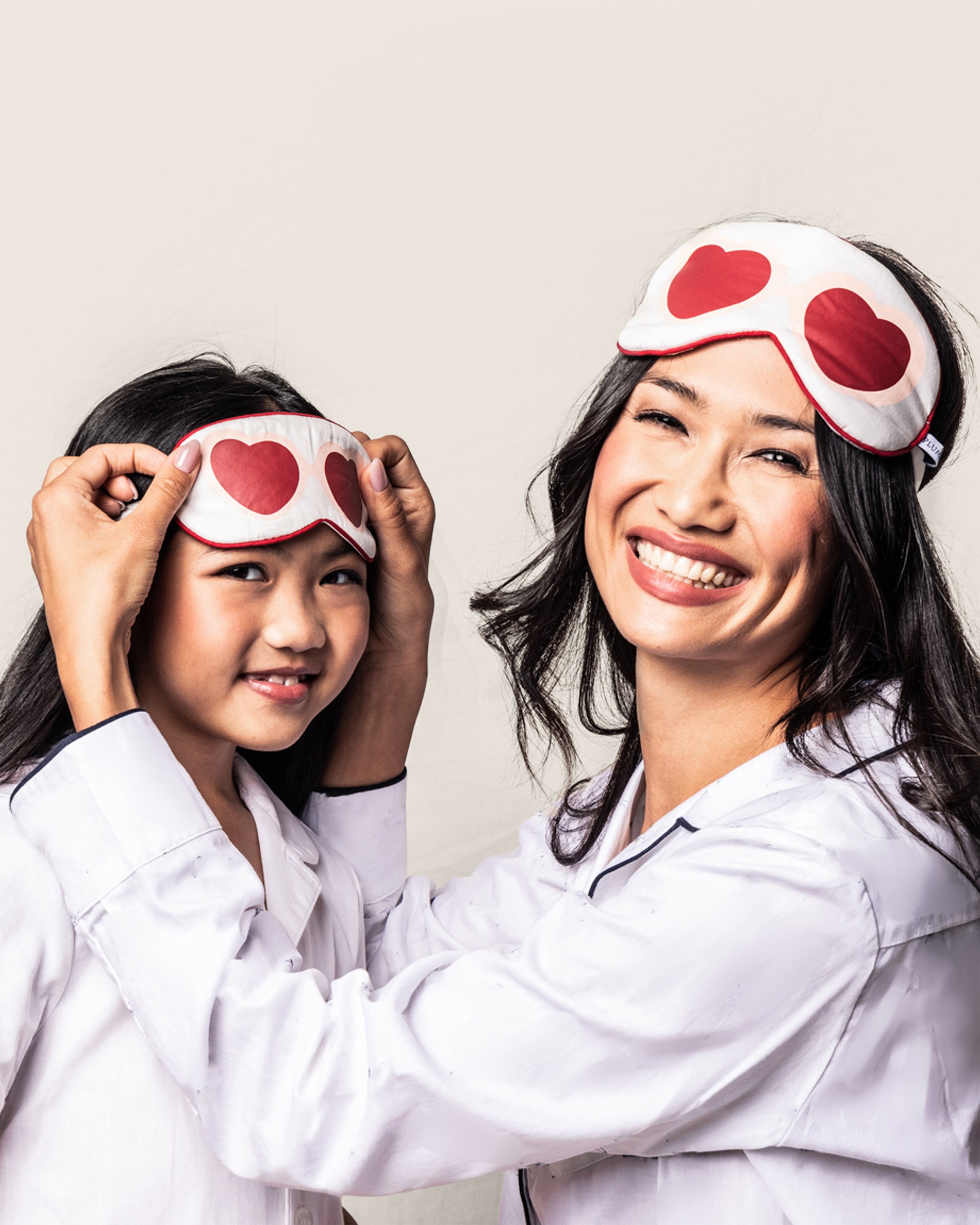 A smiling woman adjusts a Petite Plume Maisonette Exclusive Je tadore Valentines Sleep Mask on a young girls head. The girl, also smiling, wears a matching sleep mask on her forehead. They both wear luxurious sleepwear against a plain background.