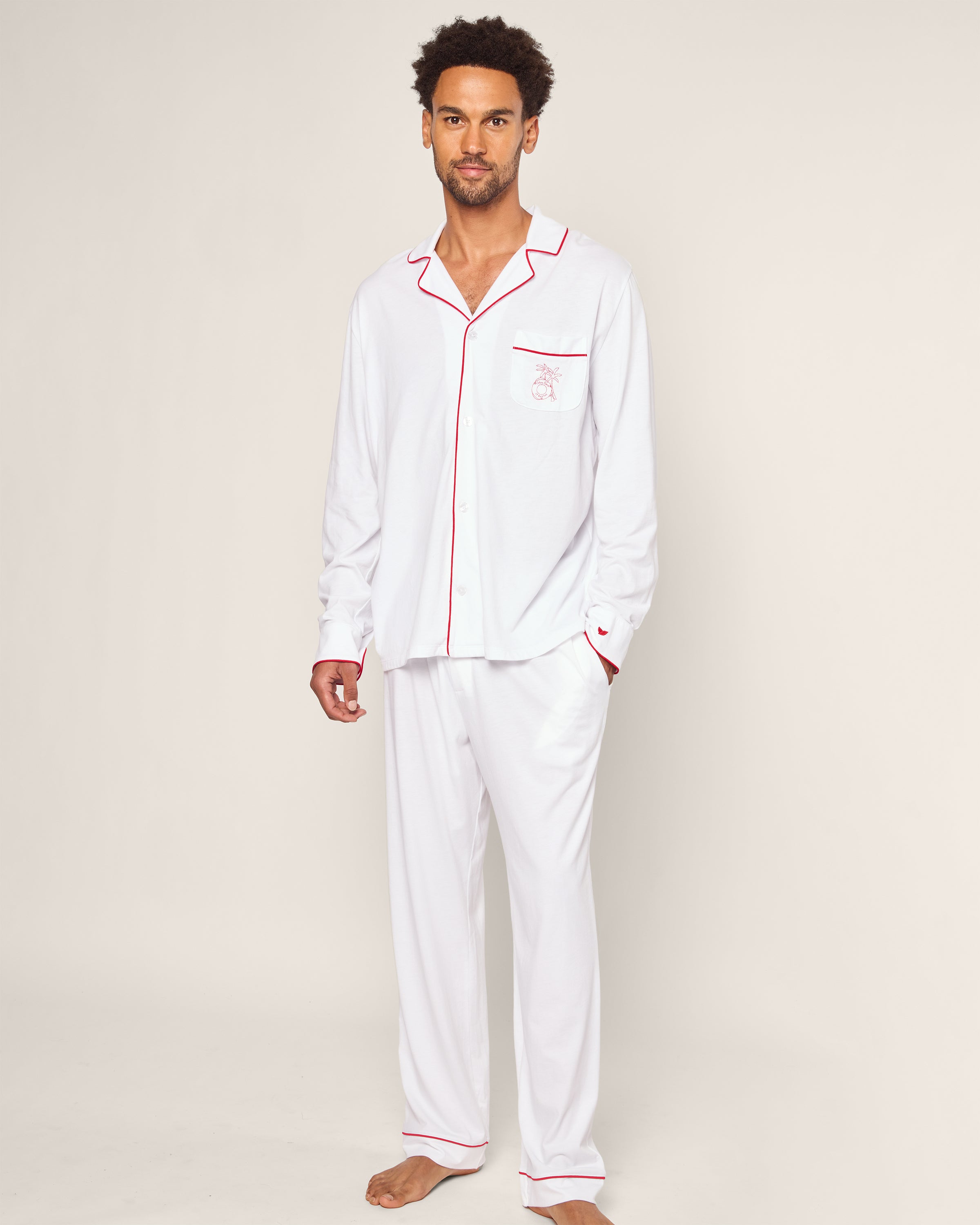A person stands barefoot in the luxurious Petite Plume x Eden Rock St Barth Mens White Pima Pajama Set. This limited-edition collection features long sleeves, a collar with red piping, and an embroidered pocket design, evoking the serene calmness of a Caribbean paradise against a light backdrop.