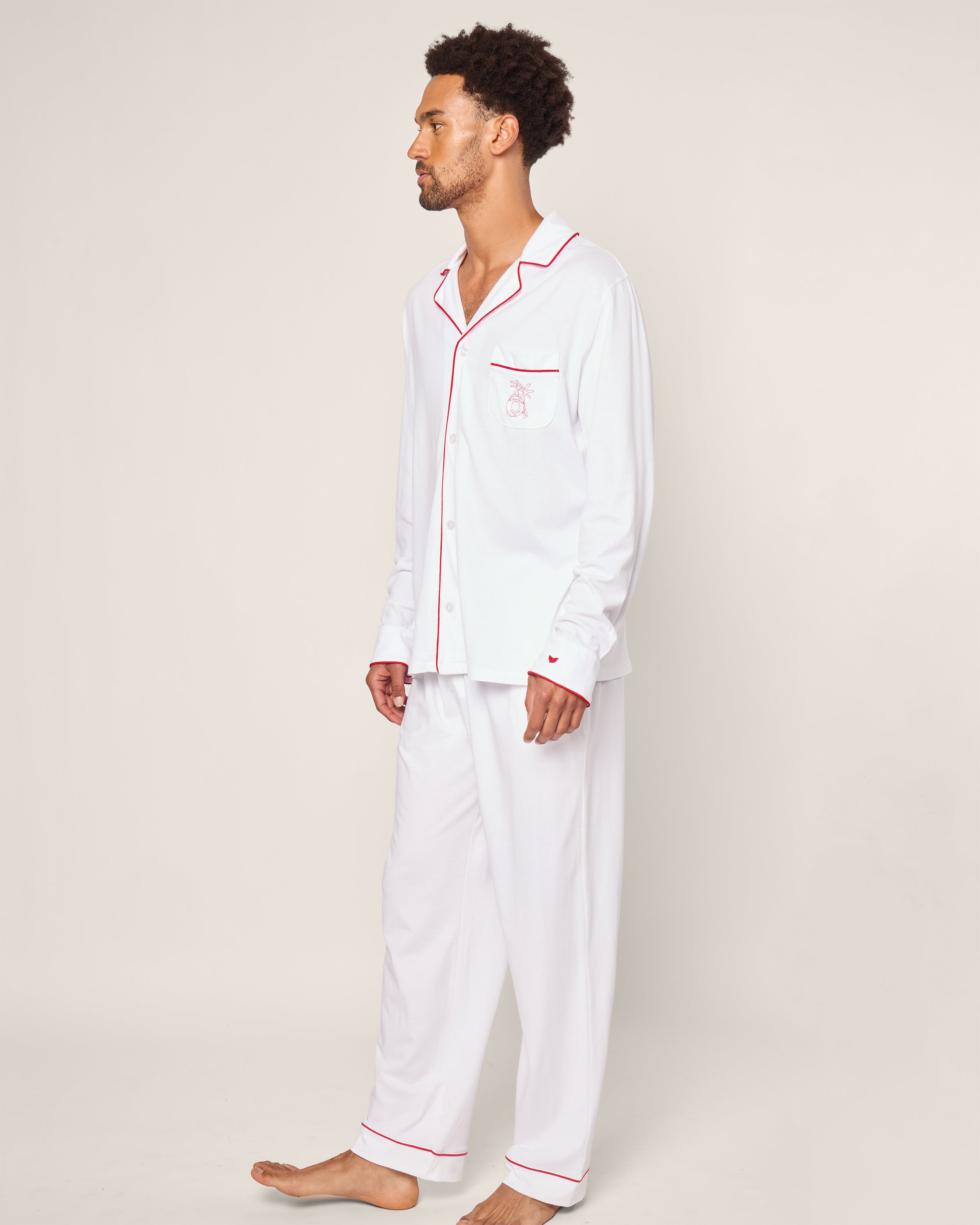 A person models the Petite Plume x Eden Rock St Barth Mens White Pima Pajama Set against a plain backdrop. This luxury sleepwear from Petite Plume includes a long-sleeve button-up shirt with a pocket and matching pants, all accented with red trim. They stand barefoot, exuding effortless elegance.