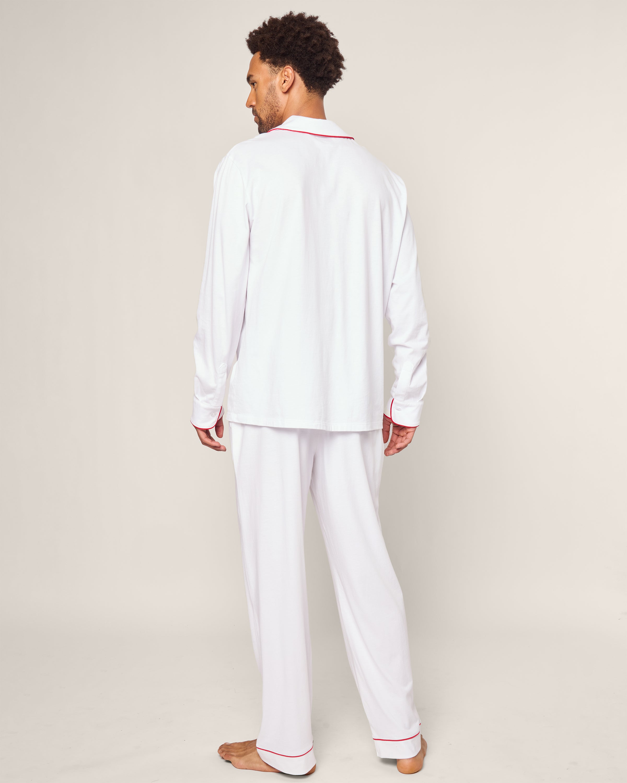 A man stands barefoot, facing away in the Petite Plume x Eden Rock St Barth Mens White Pima Pajama Set, featuring a luxury design with red trim on the collar, cuffs, and hem. The plain background highlights the elegance of this limited-edition sleepwear collection.