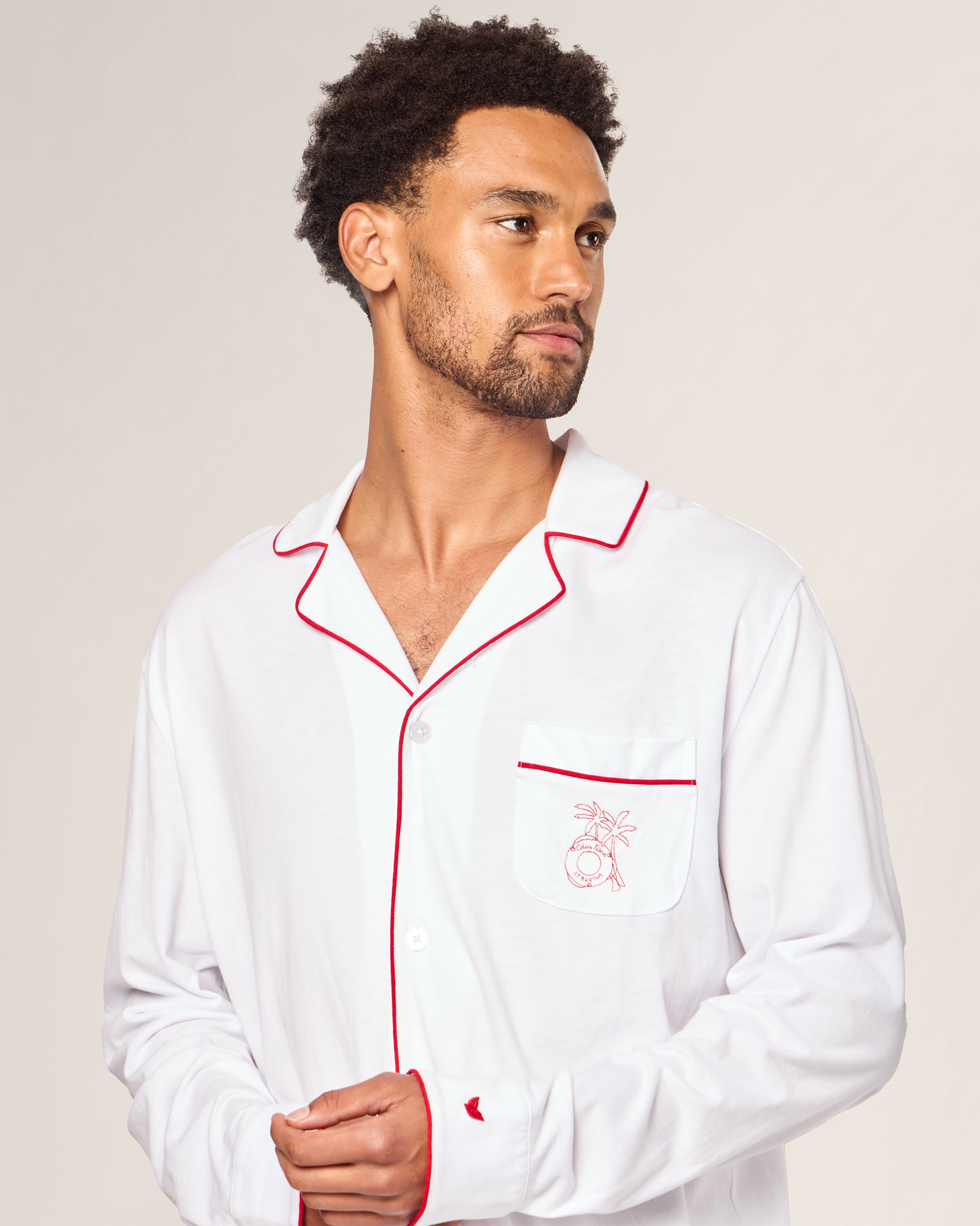 Wearing the Petite Plume x Eden Rock St Barth Mens White Pima Pajama Set, a man with short curly hair and a beard looks to the side. The luxury sleepwear, featuring red piping and an embroidered pocket design, evokes a Caribbean paradise from an exclusive limited-edition collection.
