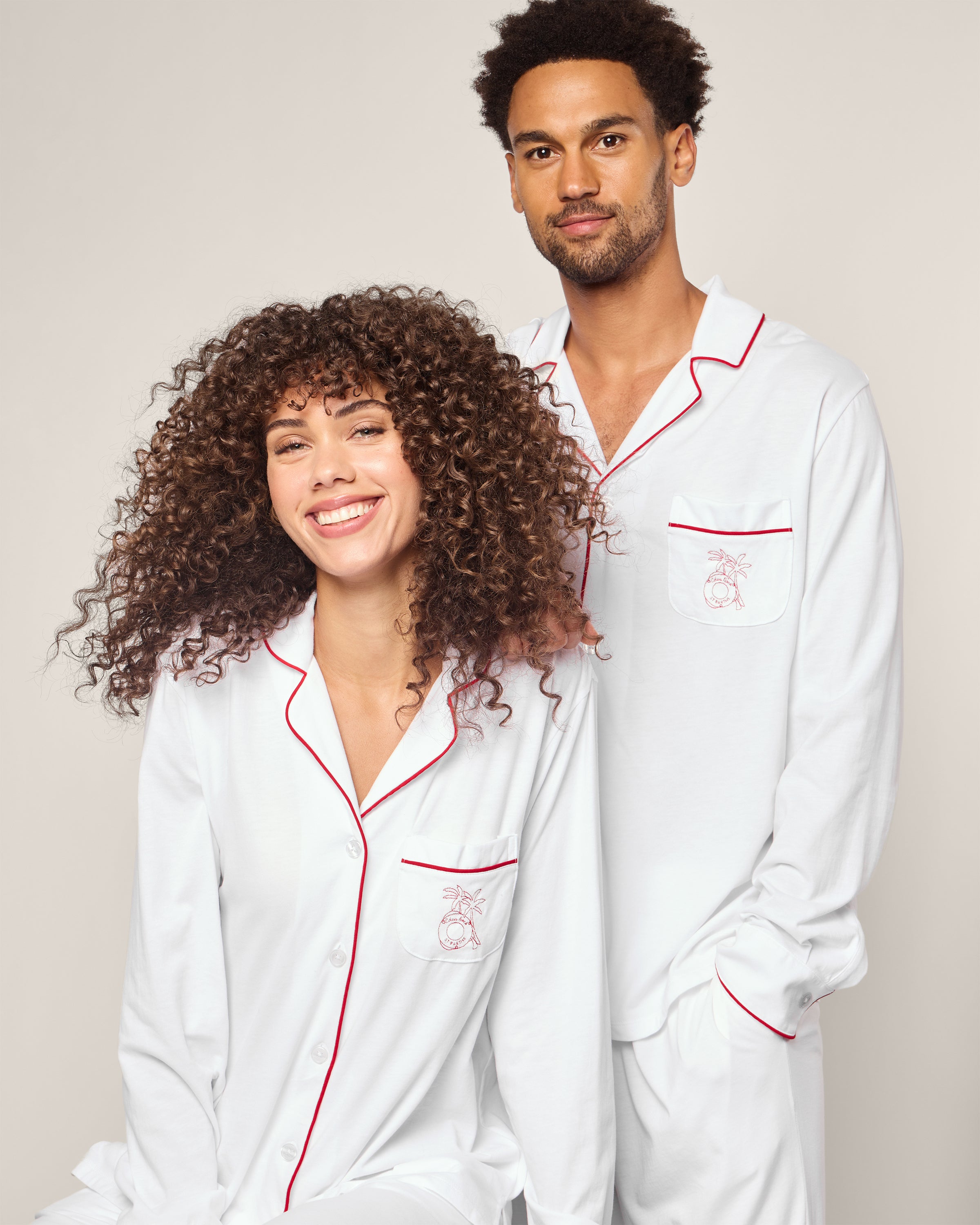A smiling woman in curly hair sits in a white pajama set with red trim, facing a man with short curly hair. Both wear luxury sleepwear from our limited-edition Petite Plume x Eden Rock St Barth collection, featuring embroidered chest pocket designs for an exclusive touch.
