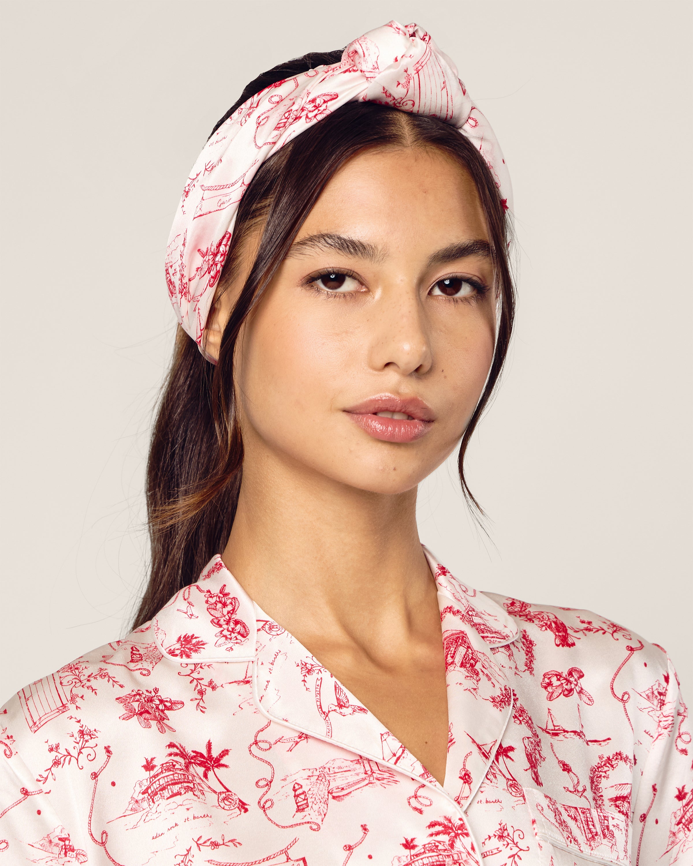A person wears a Petite Plume x Eden Rock Reverie Silk Scarf, featuring pink and red patterns from the limited-edition collection, paired with a matching shirt. Both pieces exhibit intricate designs against a plain light background that highlights the ensemble.