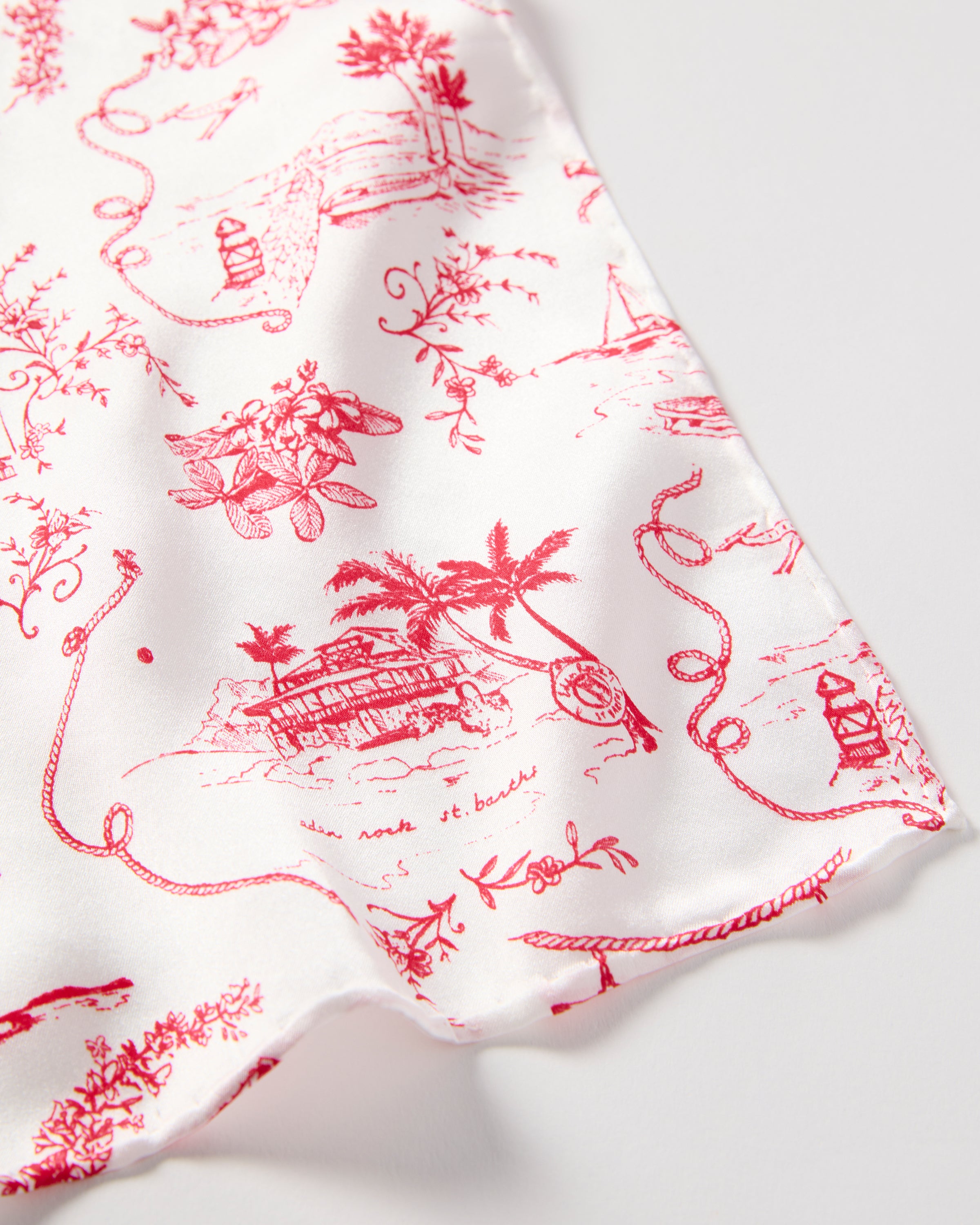 The Petite Plume x Eden Rock Reverie Silk Scarf is a white silk accessory with red toile-style illustrations, featuring intricate scenes and floral motifs, loosely rolled at the top and elegantly draped.