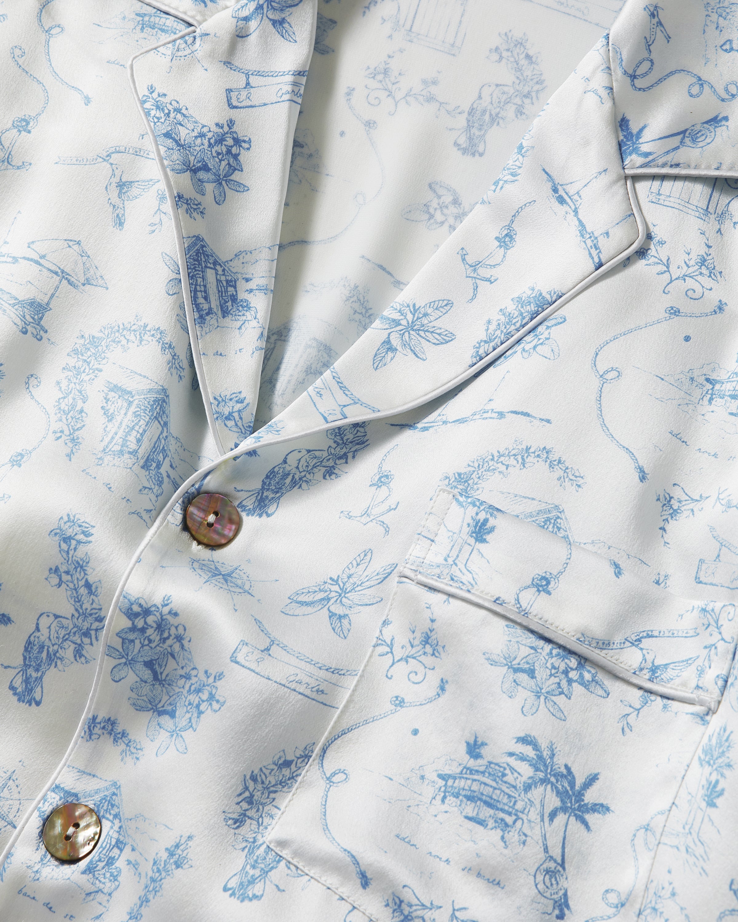 The Petite Plume x Eden Rock Blue Reverie Men's Silk Pajama Set features a white two-piece design with long sleeves and pants, light blue toile print, notched collar, front buttons on the top, and an elastic waistband on the pants.